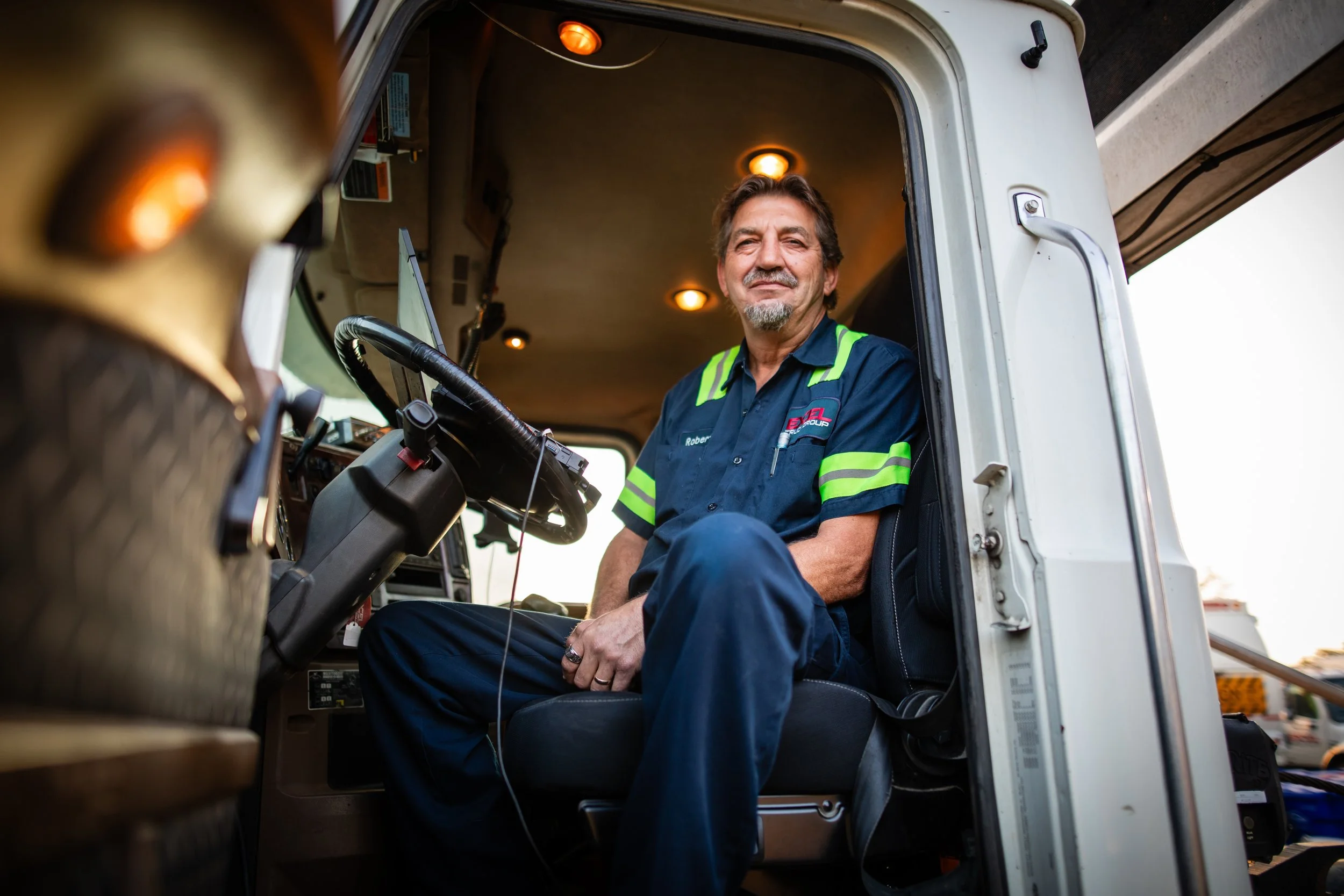 A man in a blue uniform with reflective stripes sitting inside a truck's cab, smiling at the camera.
