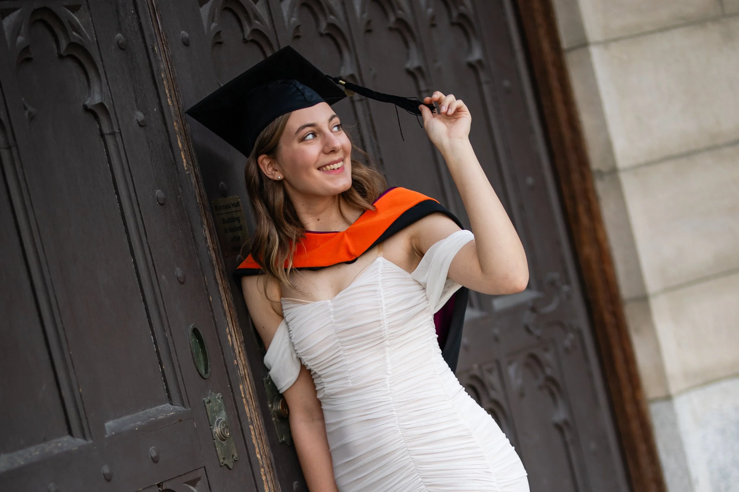 A young woman in a white dress with ruched detailing is celebrating her graduation outside a building with a large wooden door. She is wearing a black graduation cap with a tassel and an orange and black hood, and she is smiling while holding her cap