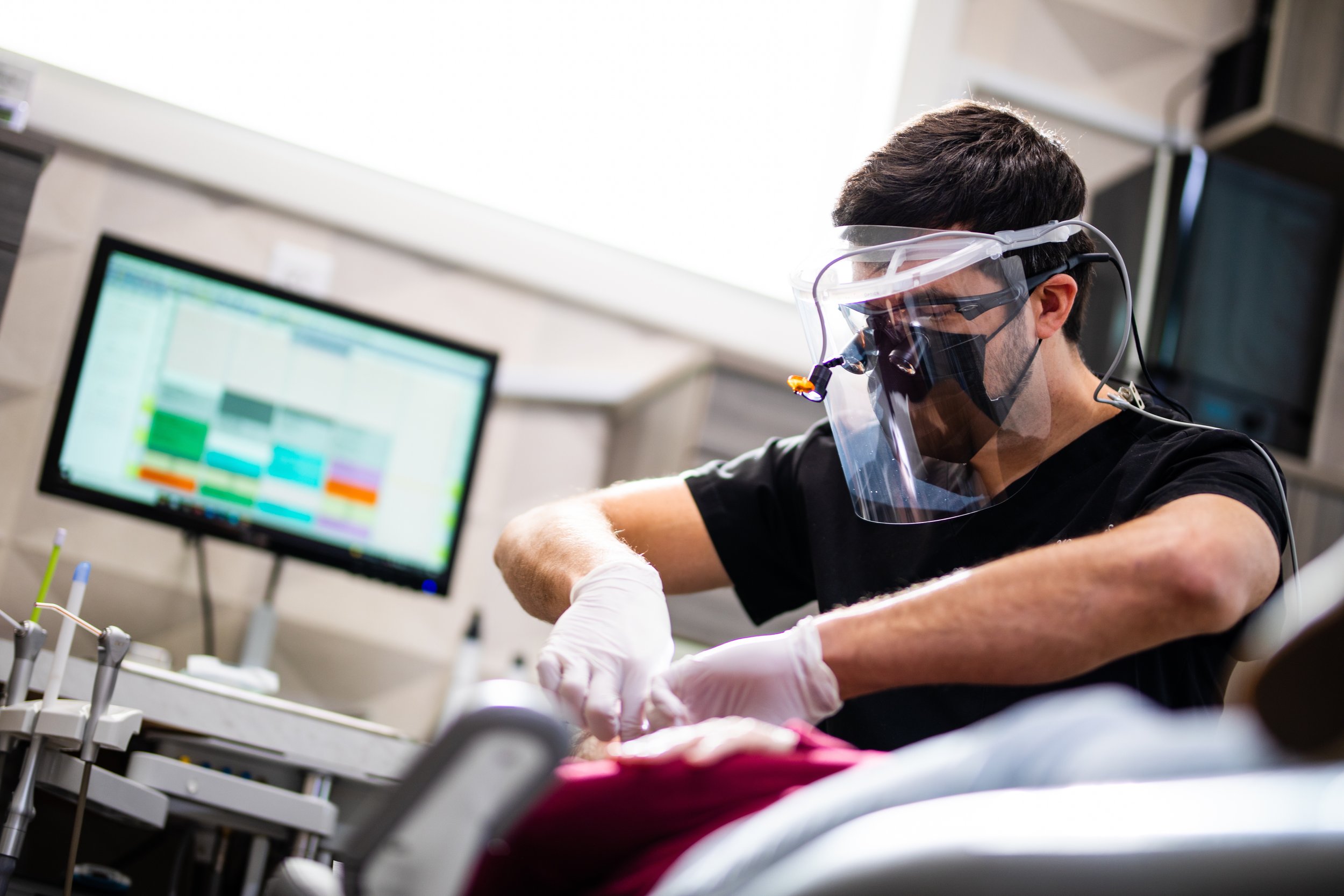 A person wearing protective gear, including a face shield and gloves, is working on a medical procedure or dental treatment in a clinical setting with a monitor in the background.