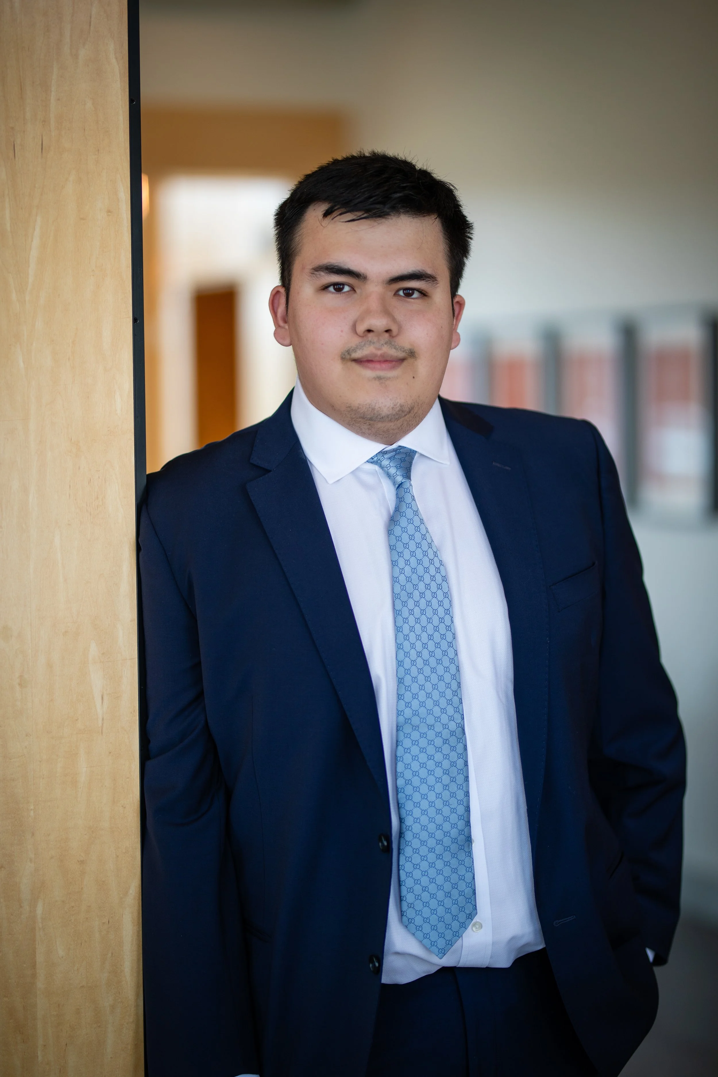 A young man with dark hair and a light mustache, wearing a navy blue suit, light blue patterned tie, and white dress shirt, standing indoors against a wooden pillar with a blurred hallway background.