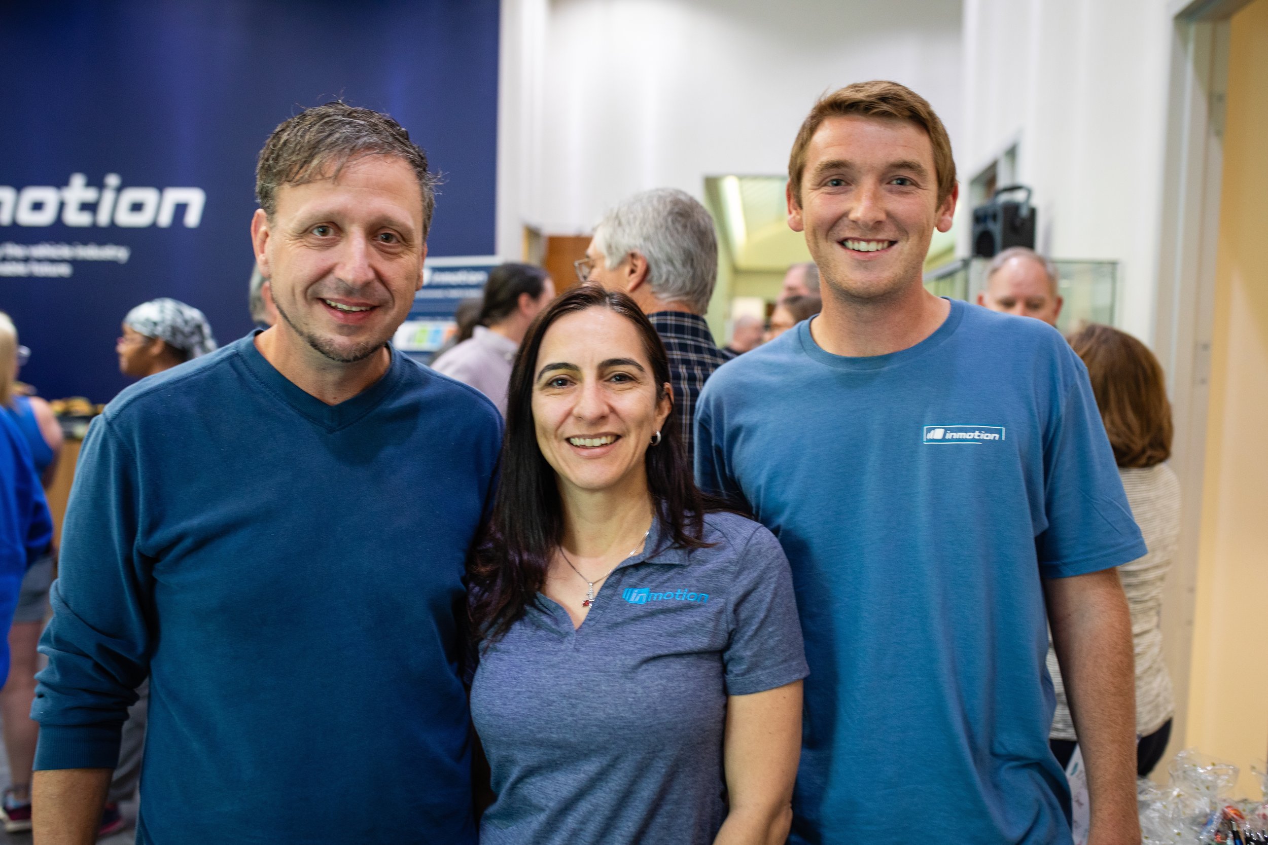 Three smiling people standing together at an event, with a crowd and a blue wall with the logo 'inmotion' in the background.