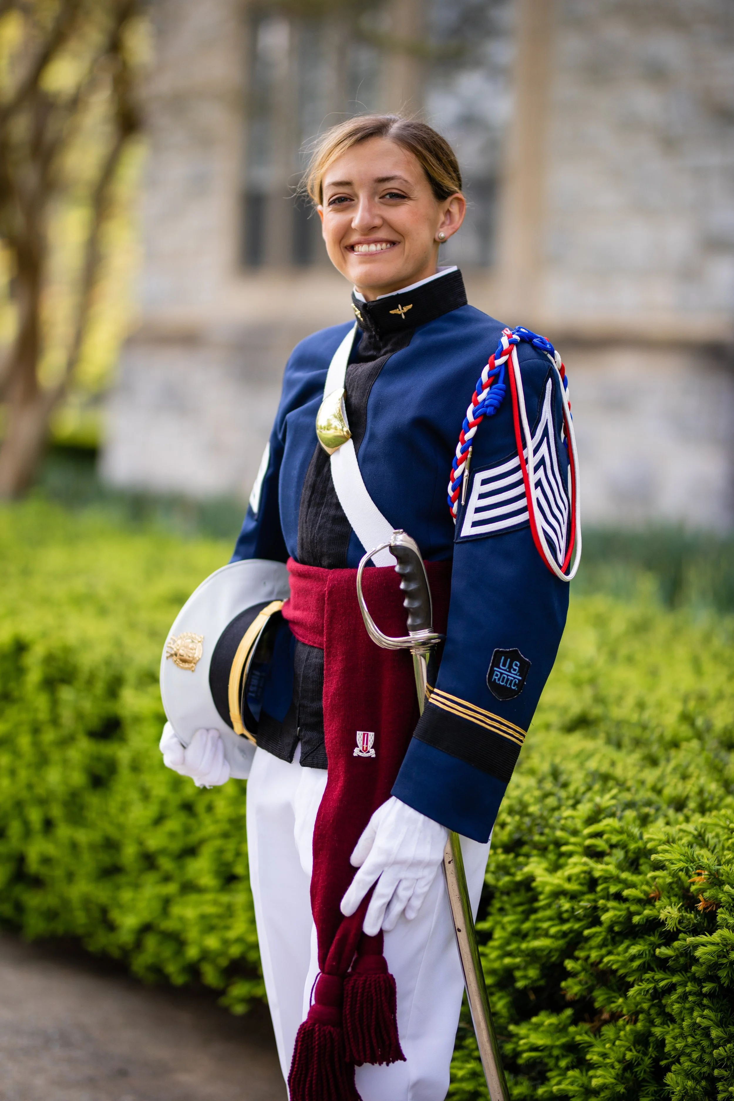 A young woman in a military marching band uniform holding a white hat and a red plume, standing outdoors with greenery and a stone building in the background, smiling.