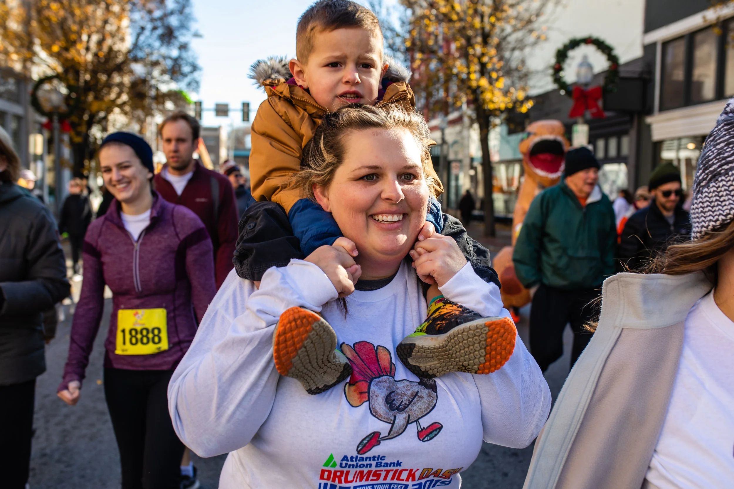 A woman with a child on her shoulders during a festive outdoor event. The woman is smiling, and the child appears to be looking worried. People are walking in the background, with some wearing race bibs and one dressed in a dinosaur costume. The scen