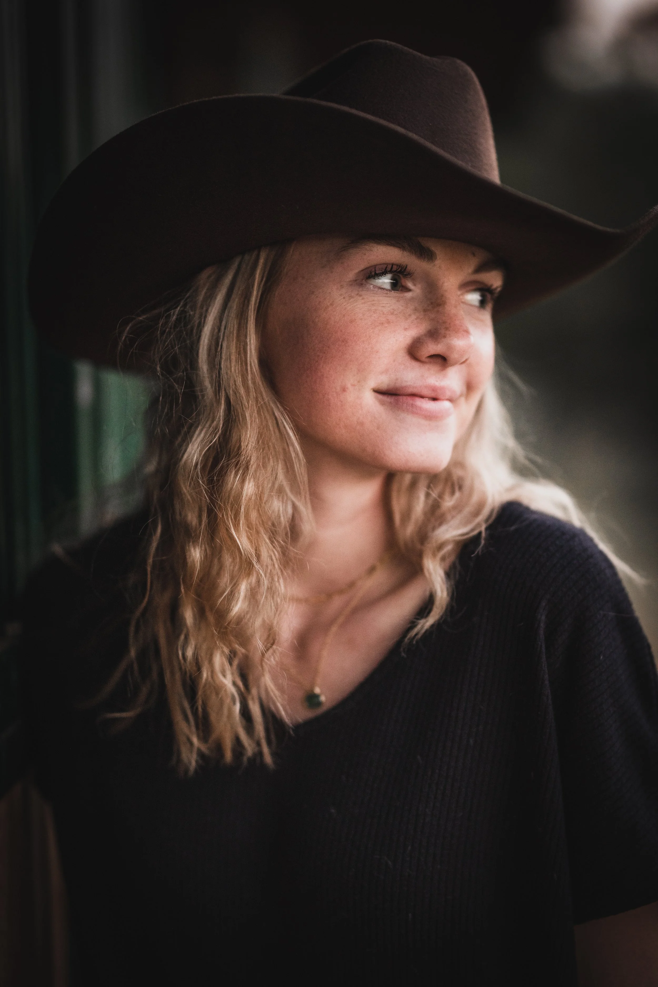 A woman with wavy blonde hair wearing a large brown cowboy hat and a black top, looking away with a gentle smile.