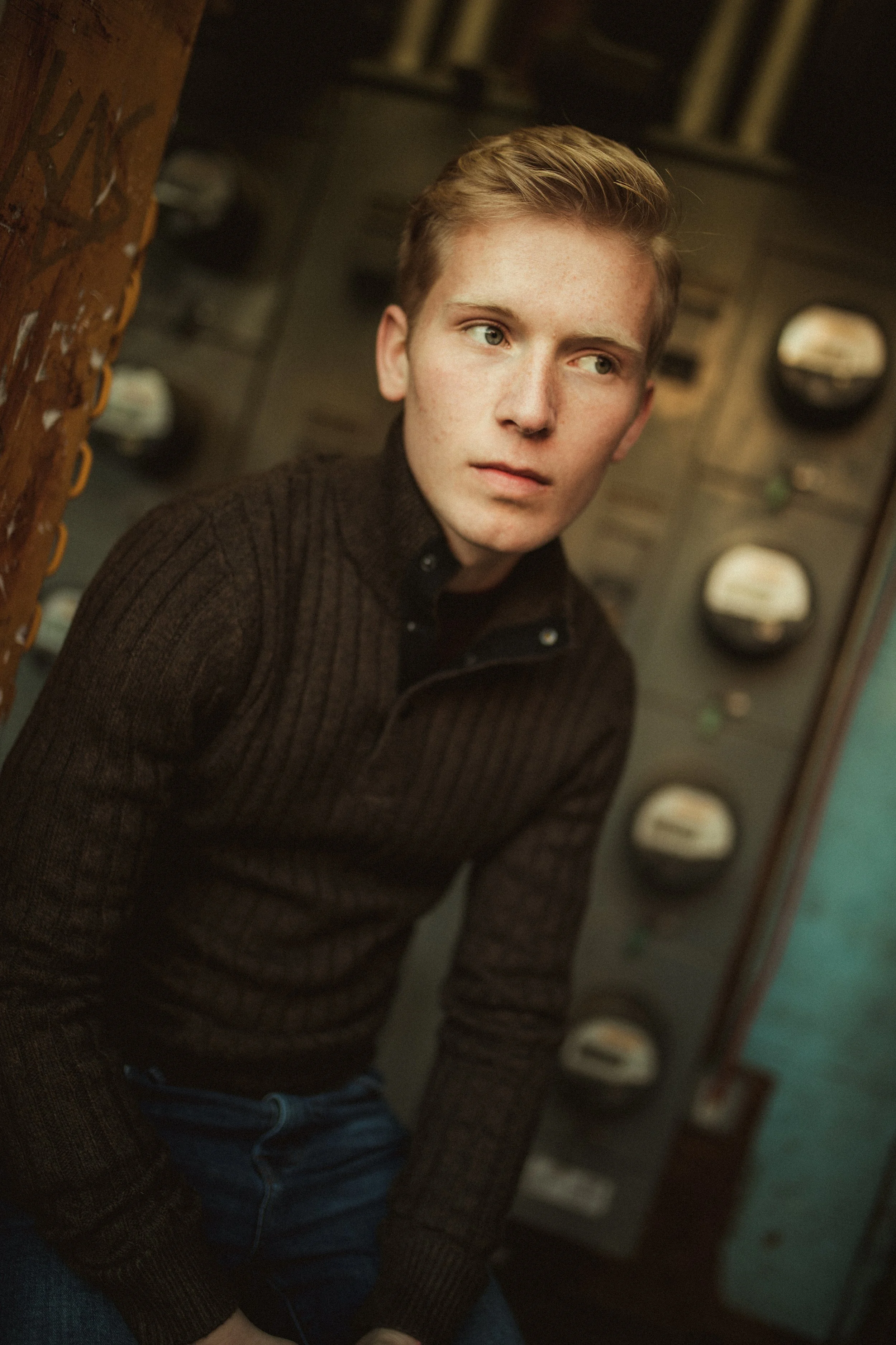 A young man with blond hair, wearing a dark brown knit sweater, is sitting near a control panel with several round meters or gauges. He is looking off to the side with a contemplative expression.