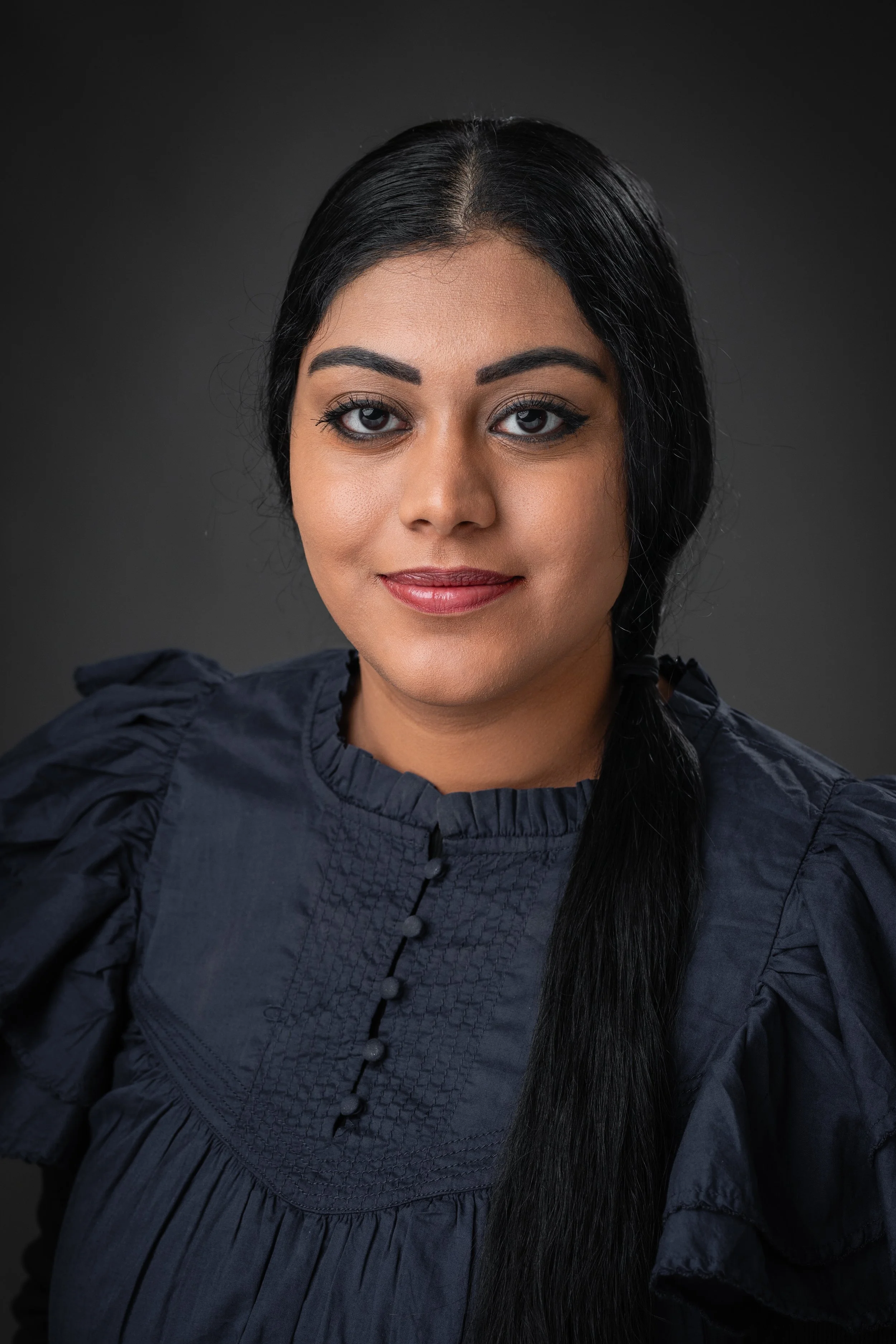 Close-up portrait of a woman with dark hair, makeup, wearing a dark blouse with buttoned details, against a dark background.