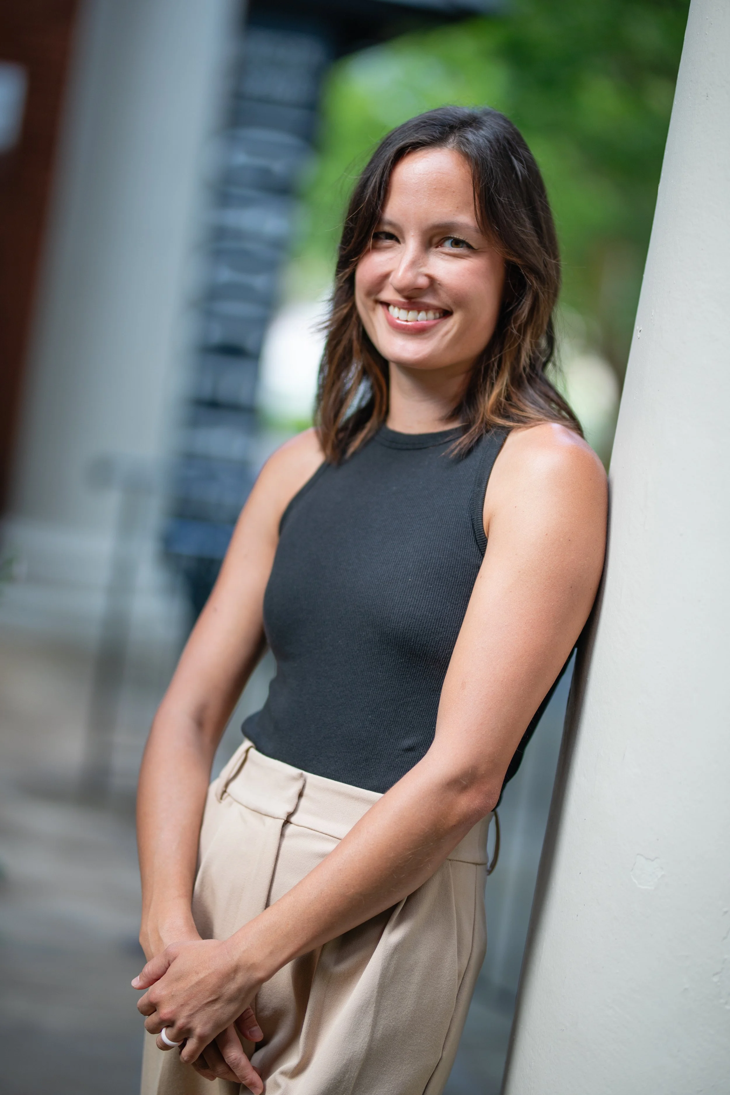 Young woman smiling, leaning against a wall outdoors, wearing a black sleeveless top and beige pants.