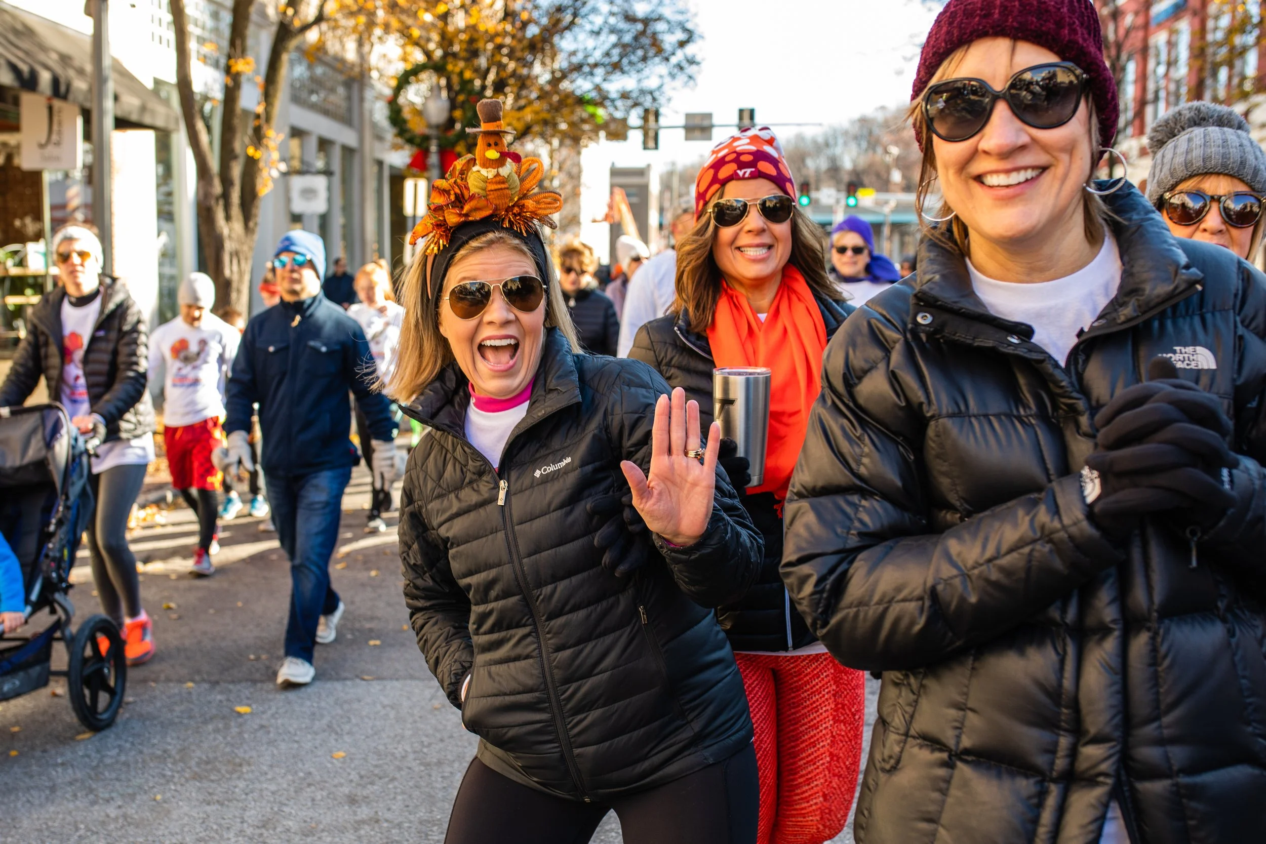 Women smiling, wearing sunglasses and winter jackets, walking outdoors during a festive event or marathon, autumn trees in the background.