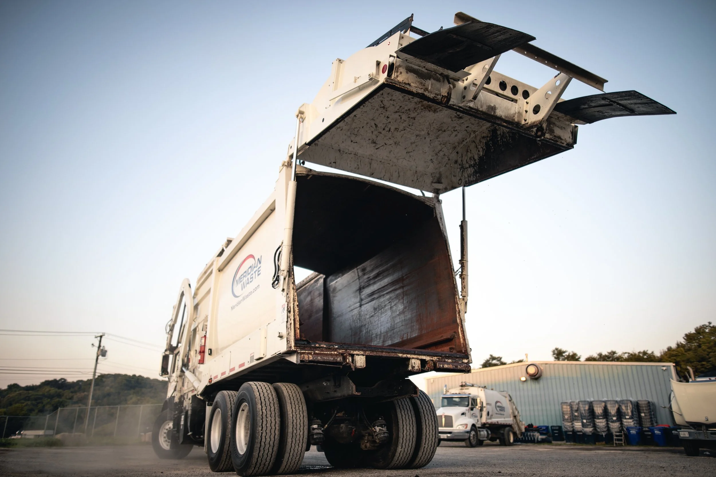 A large medical waste truck with its back open, showing an empty storage compartment. The truck is white with a logo that says 'Medical Waste'. The photo is taken outdoors at a facility with other trucks and equipment in the background.