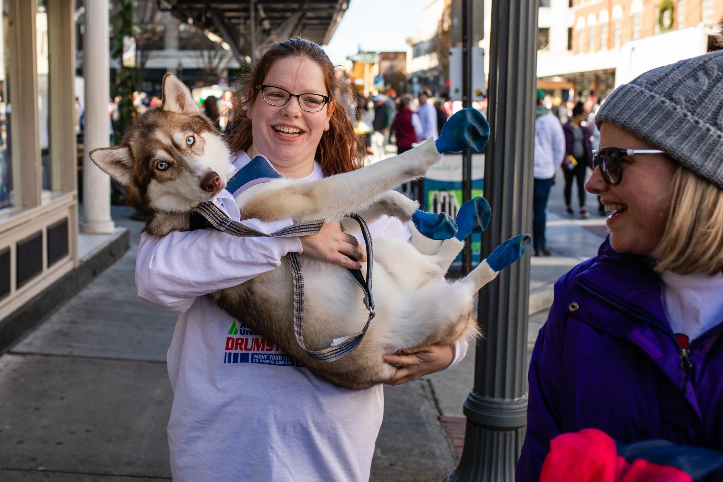 A young woman holding a Siberian Husky puppy with blue eyes, who is wearing blue gloves. A woman wearing sunglasses and a beanie is smiling at the young woman and puppy, in an outdoor street setting with people in the background.