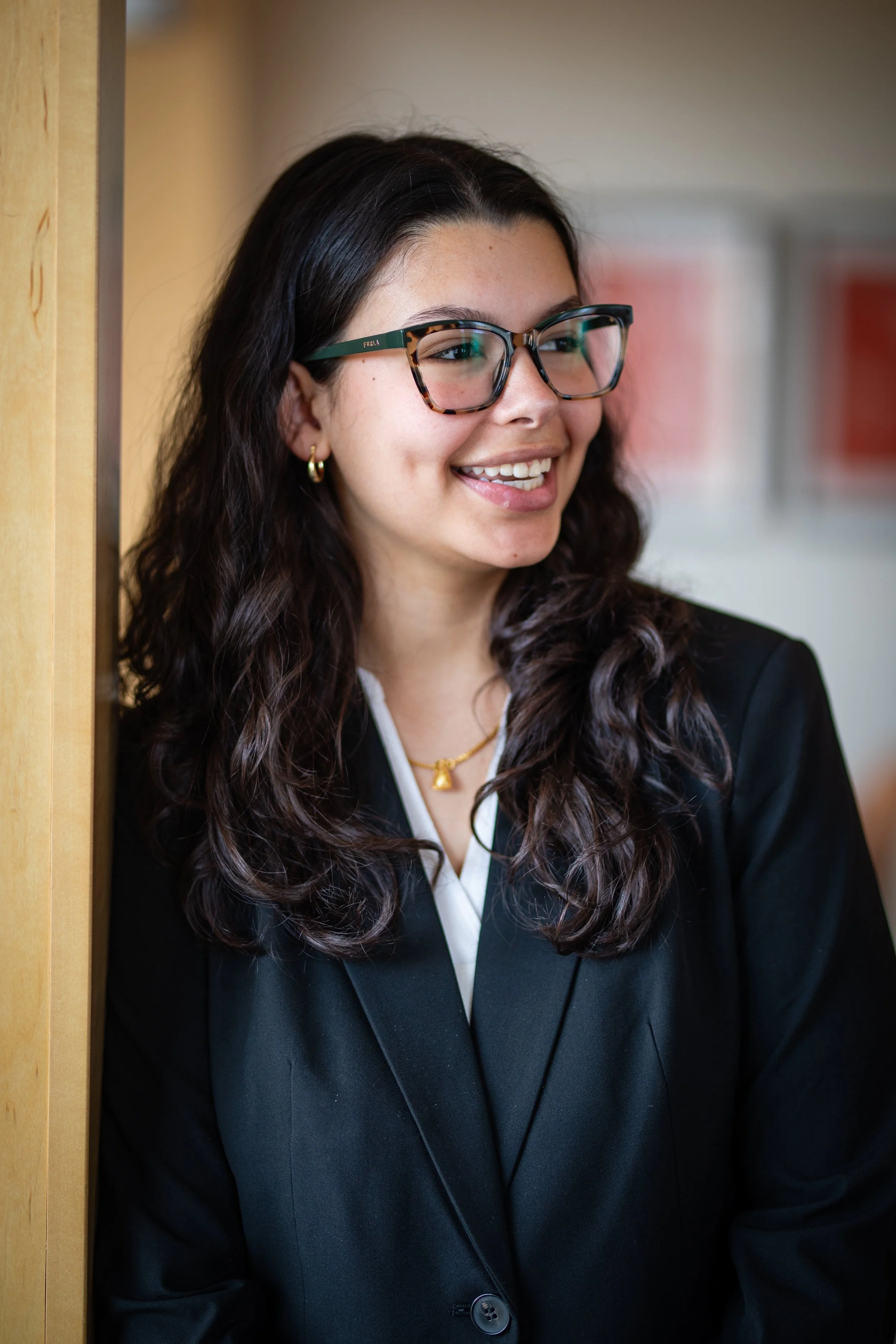 A woman with long dark curly hair, glasses, and gold jewelry, wearing a black blazer, smiling and leaning against a wooden wall indoors.