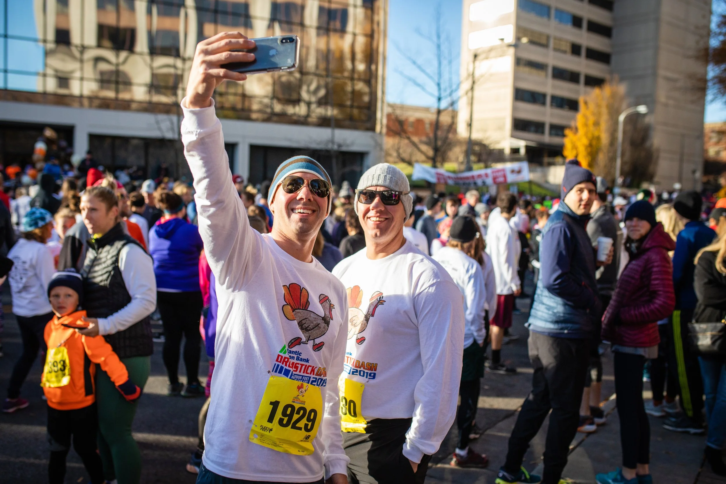 Two men in white shirts taking a selfie at an outdoor marathon event, surrounded by a crowd of runners and spectators with tall buildings in the background.
