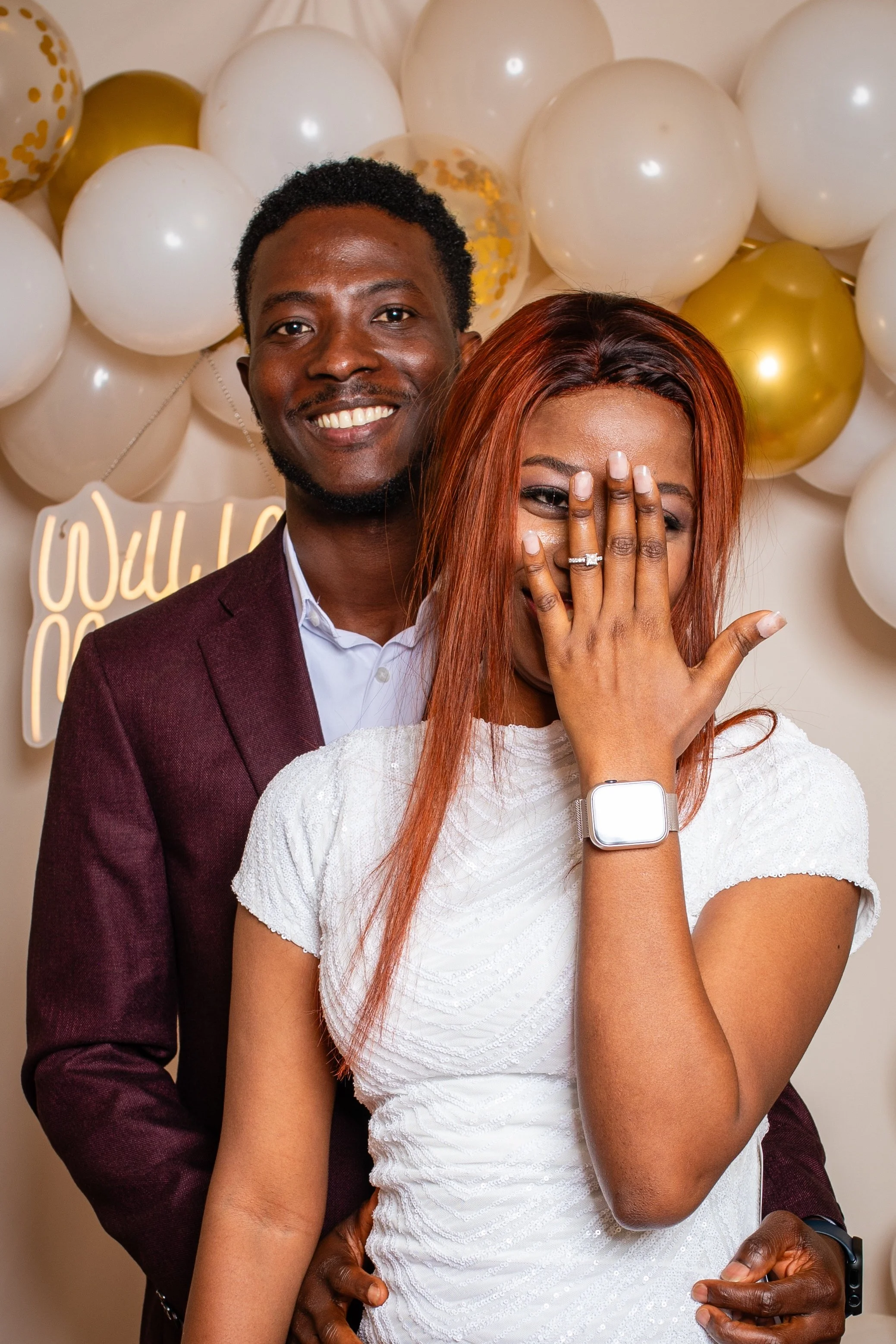 A couple standing in front of a balloon decoration, with the man smiling and the woman covering her face with her hand, showing an engagement ring.