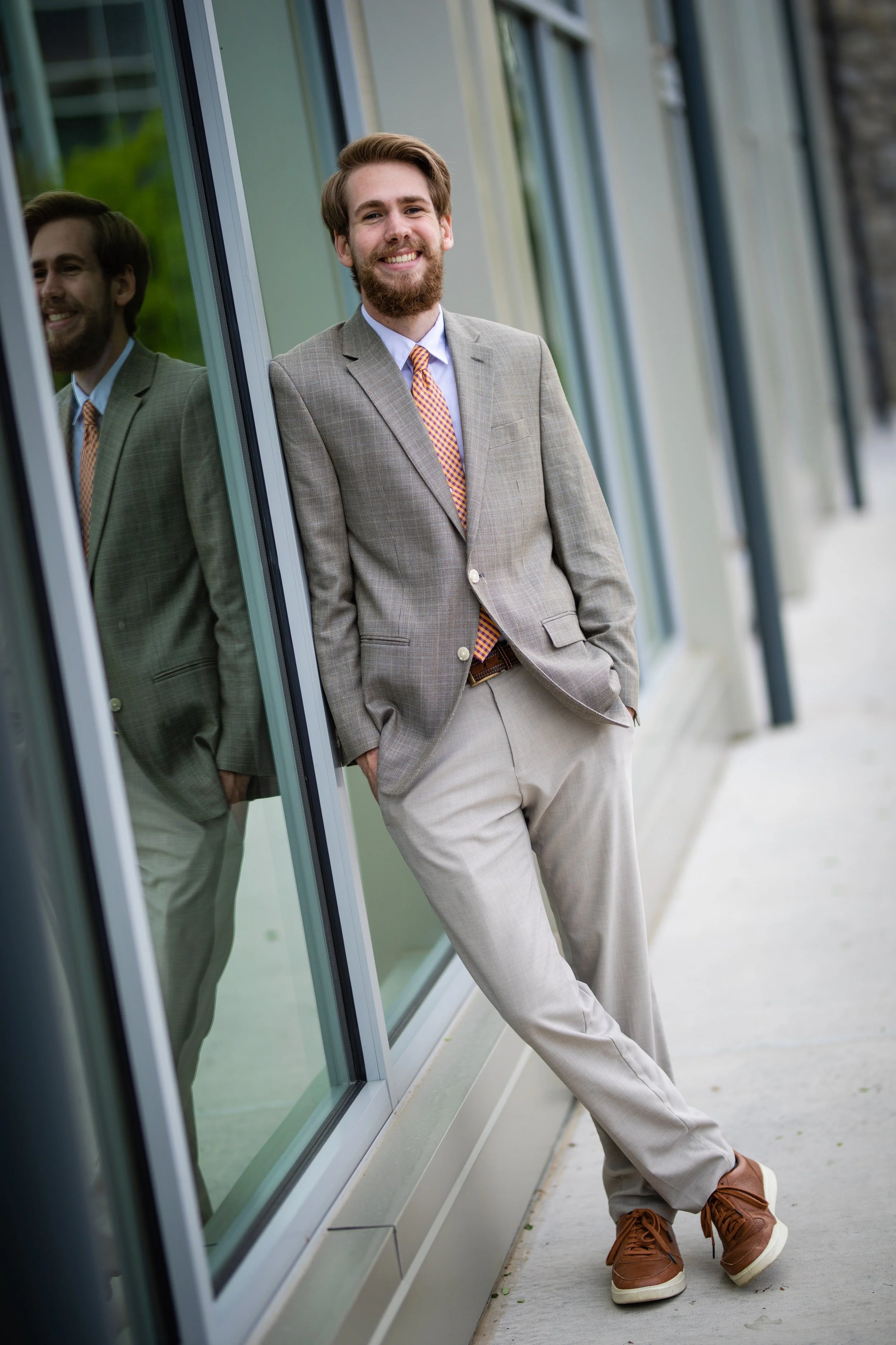 A smiling man in a light gray suit, orange checkered tie, and brown shoes standing outside a building with glass windows, leaning against the window frame with his hands in his pockets.