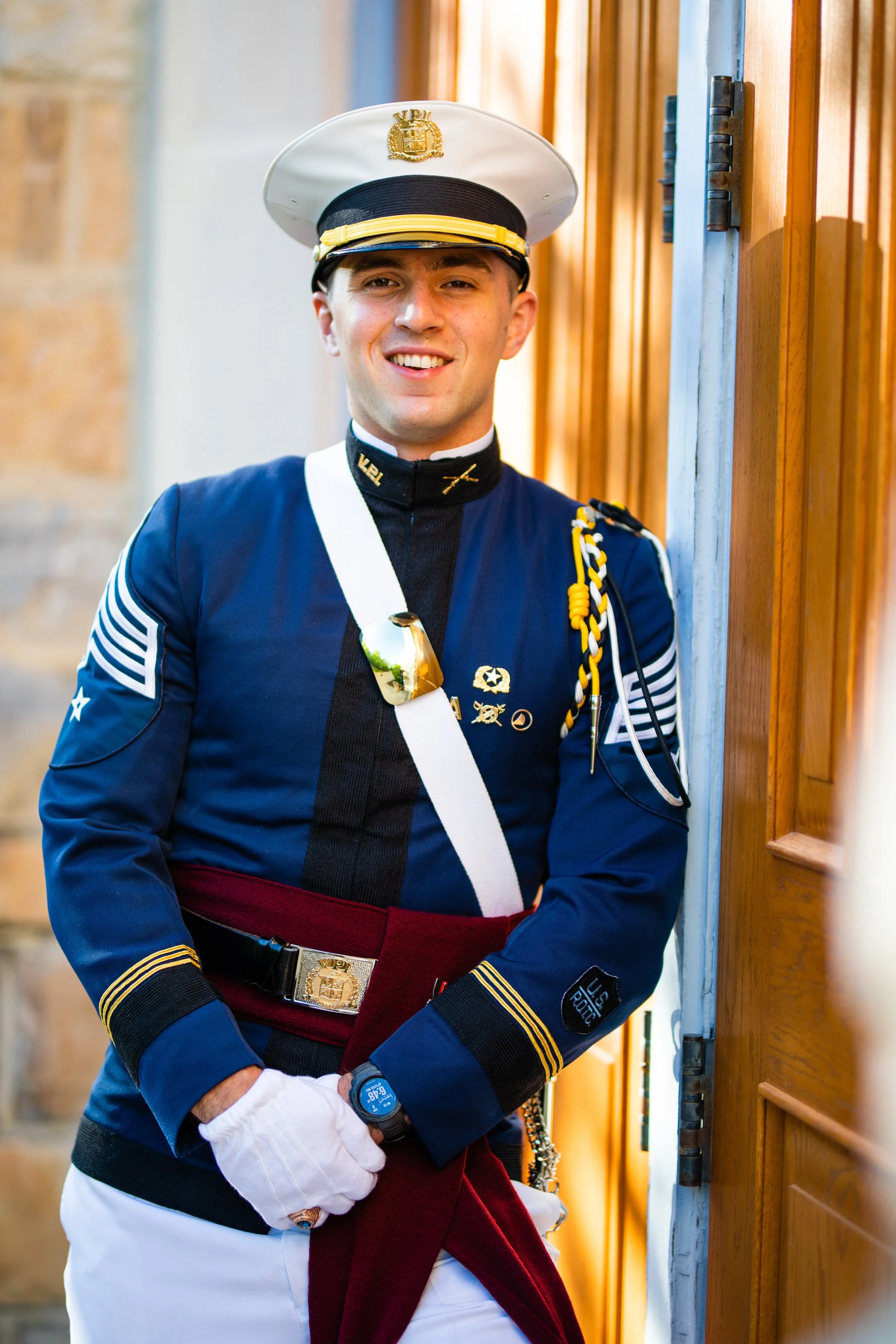 Young man in a military uniform and hat standing next to a wooden door, smiling at the camera.