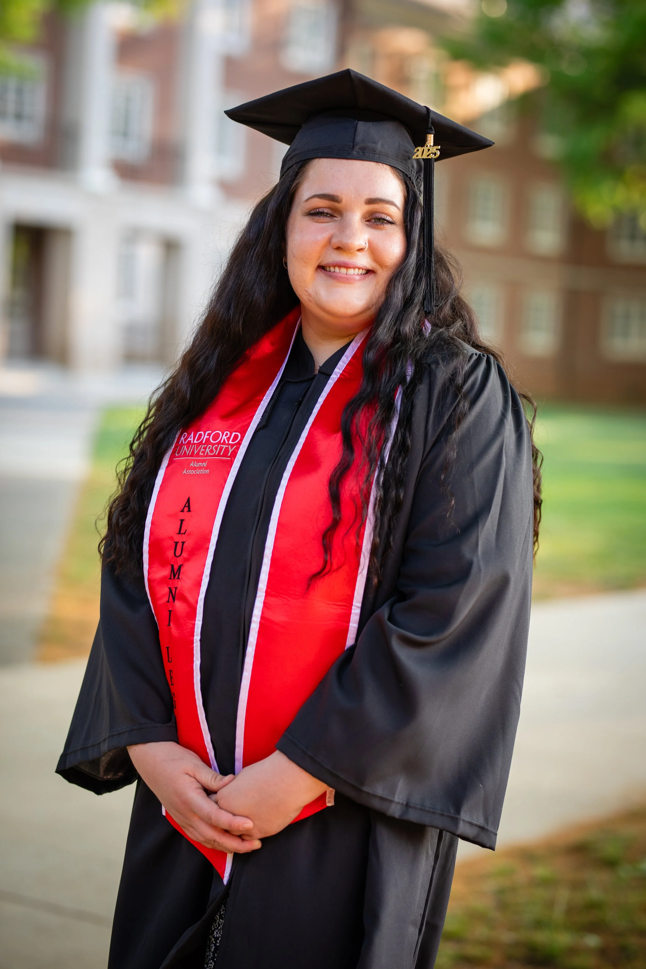 A young woman in a black graduation gown and cap, with a red stole that reads 'Alumni' and 'Radford University.' She is smiling, standing outside in front of a blurred background of buildings and trees.