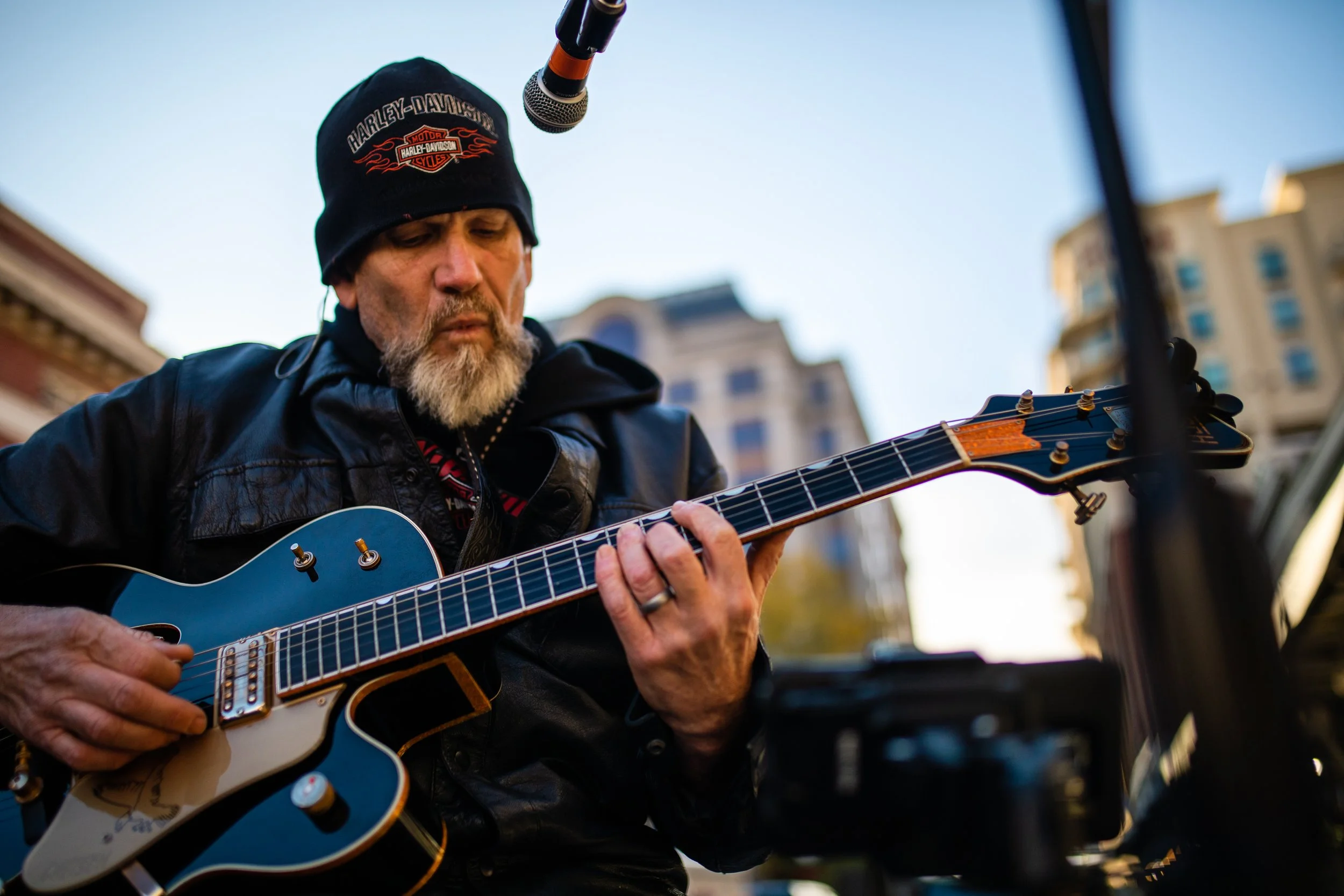 A man with a gray beard wearing a black Harley-Davidson beanie and leather jacket playing an electric guitar outdoors with a cityscape background and a microphone overhead.
