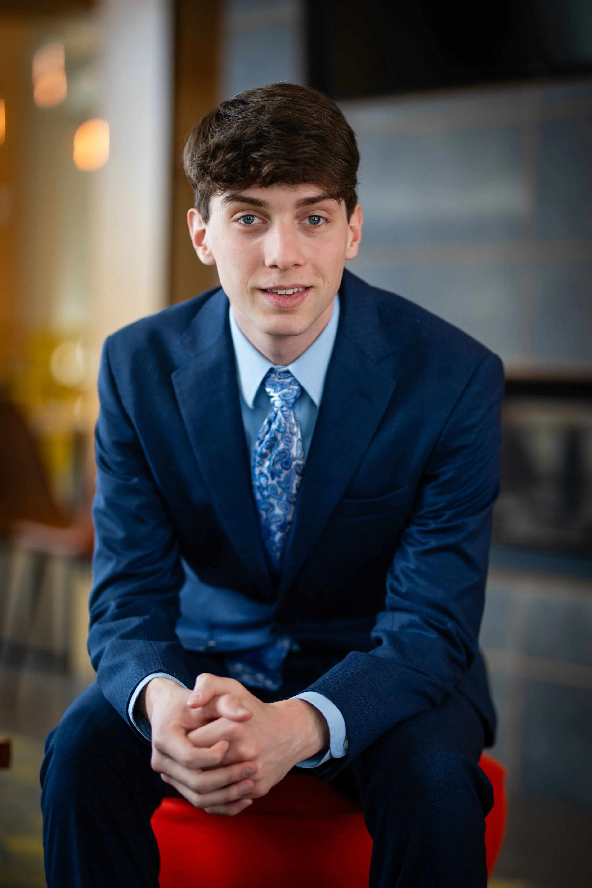 A young man in a blue suit and patterned tie, sitting and smiling, in a modern indoor setting.