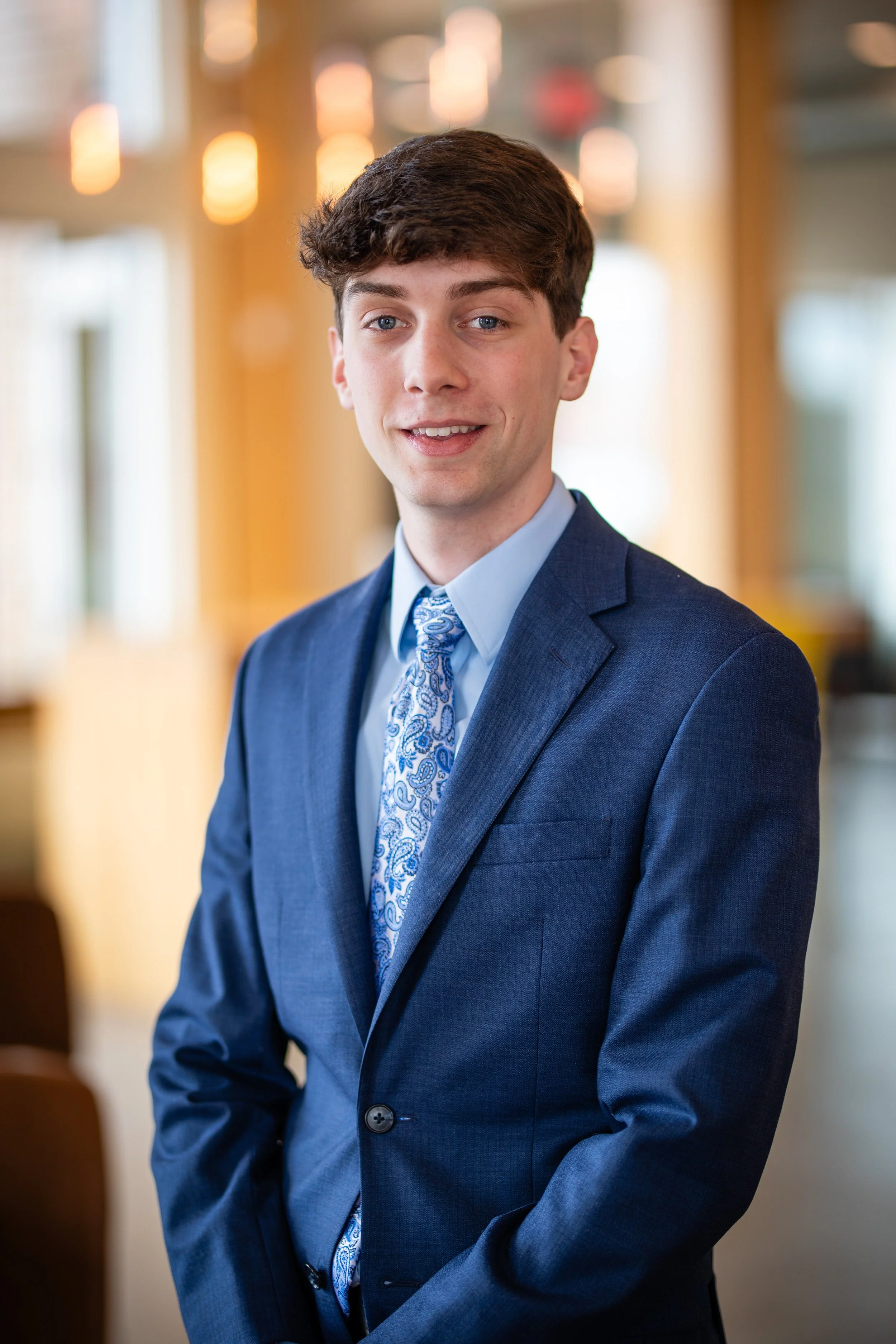 Young man in a blue suit and patterned tie standing indoors with blurred warm lighting in the background.