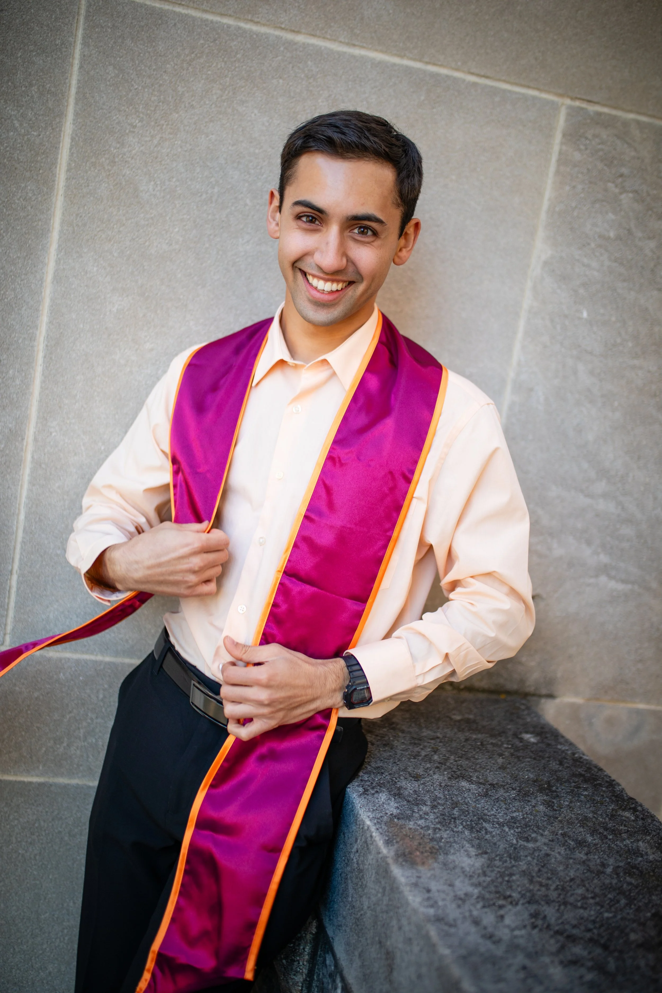 A young man smiling, wearing a peach dress shirt and black trousers, with a purple graduation stole with orange trim around his neck, standing outdoors against a stone wall.
