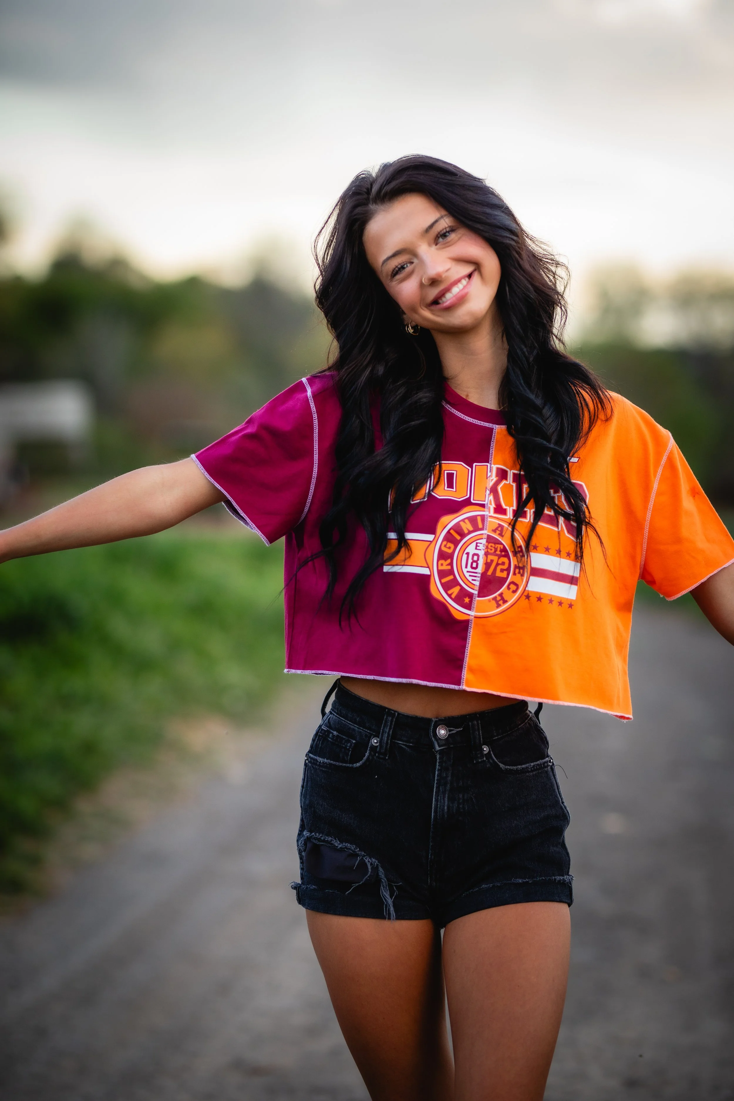 A young woman with long dark hair is smiling and standing outdoors on a path, wearing a colorful, half-oclock shirt with orange and purple sections and black shorts.