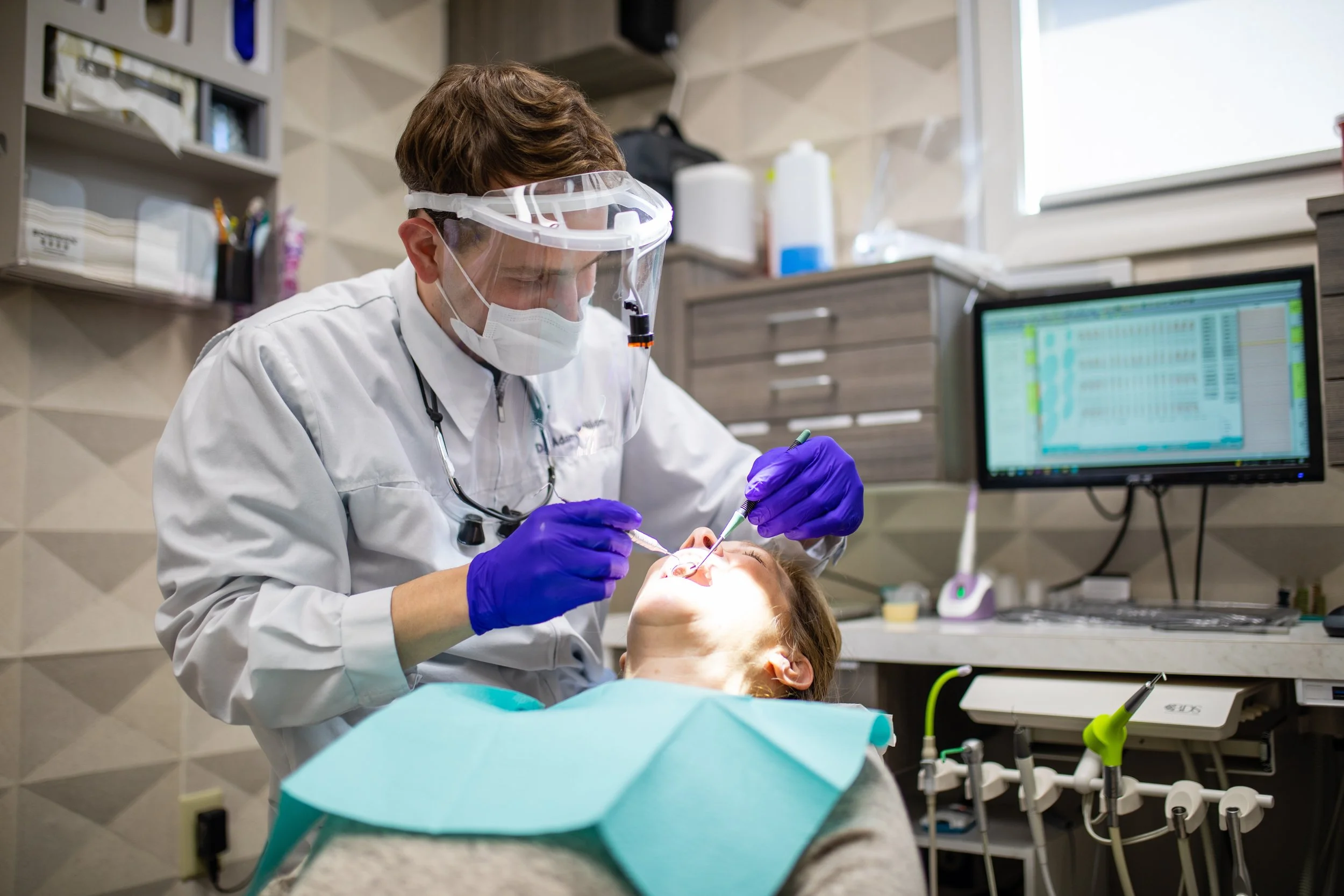 Dentist performing a dental procedure on a patient in a dental office, with dental tools, computer monitor, and dental equipment visible.