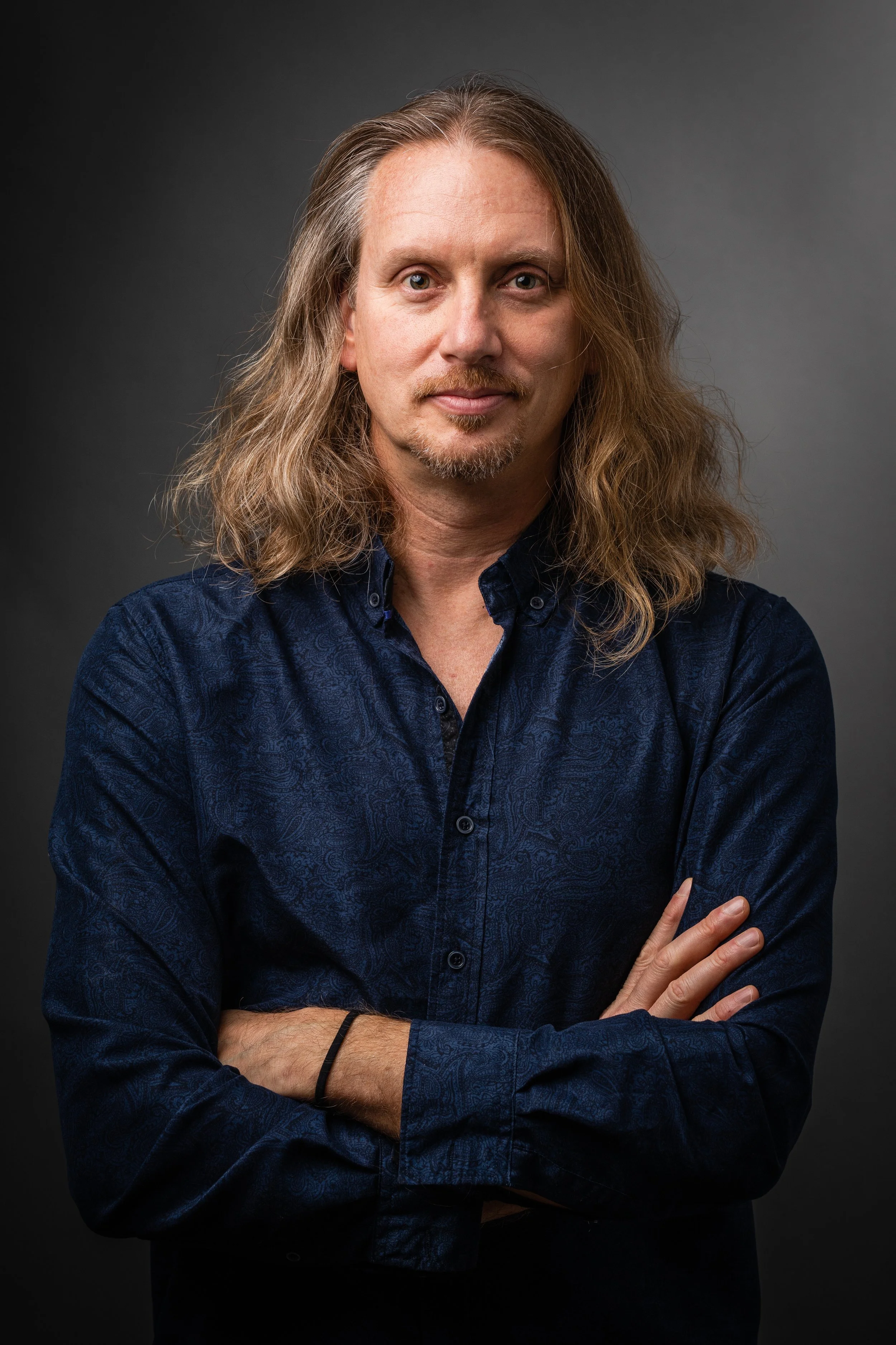 A man with long wavy hair, light skin, and a goatee, wearing a dark blue patterned shirt, standing with arms crossed against a gray background.