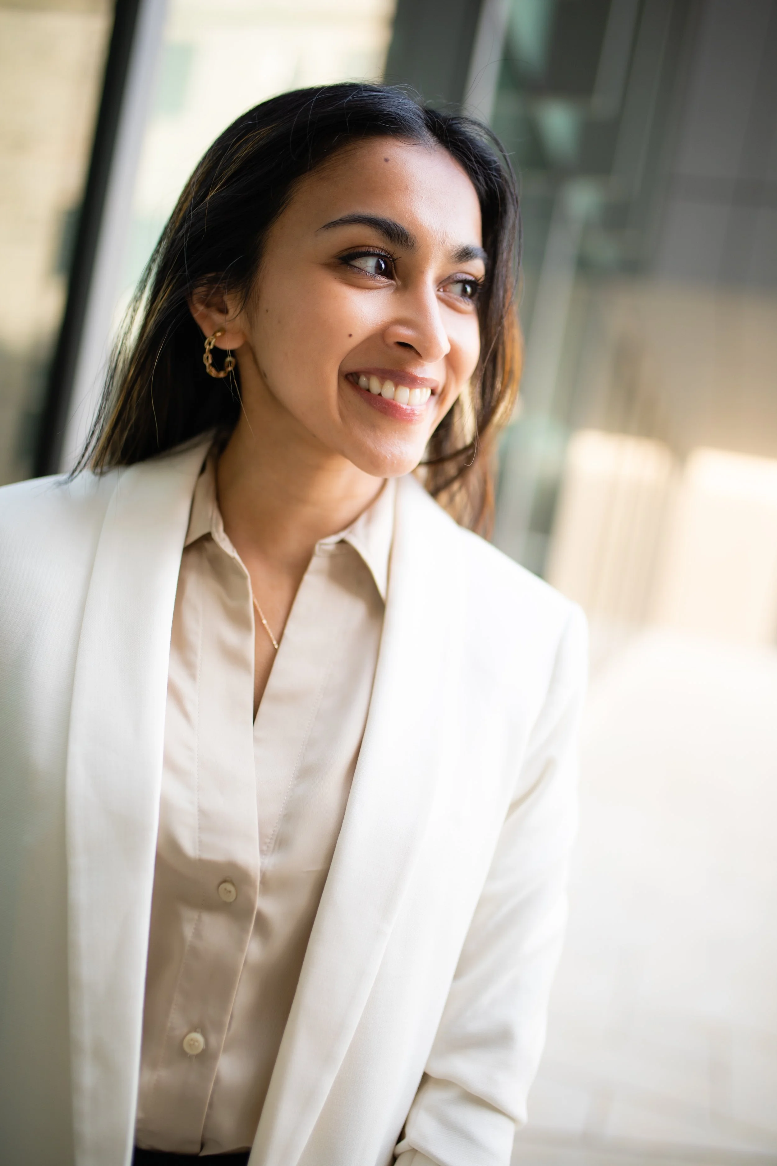A smiling woman with long dark hair and gold hoop earrings, dressed in a light-colored blazer and blouse, standing near large windows with natural sunlight.