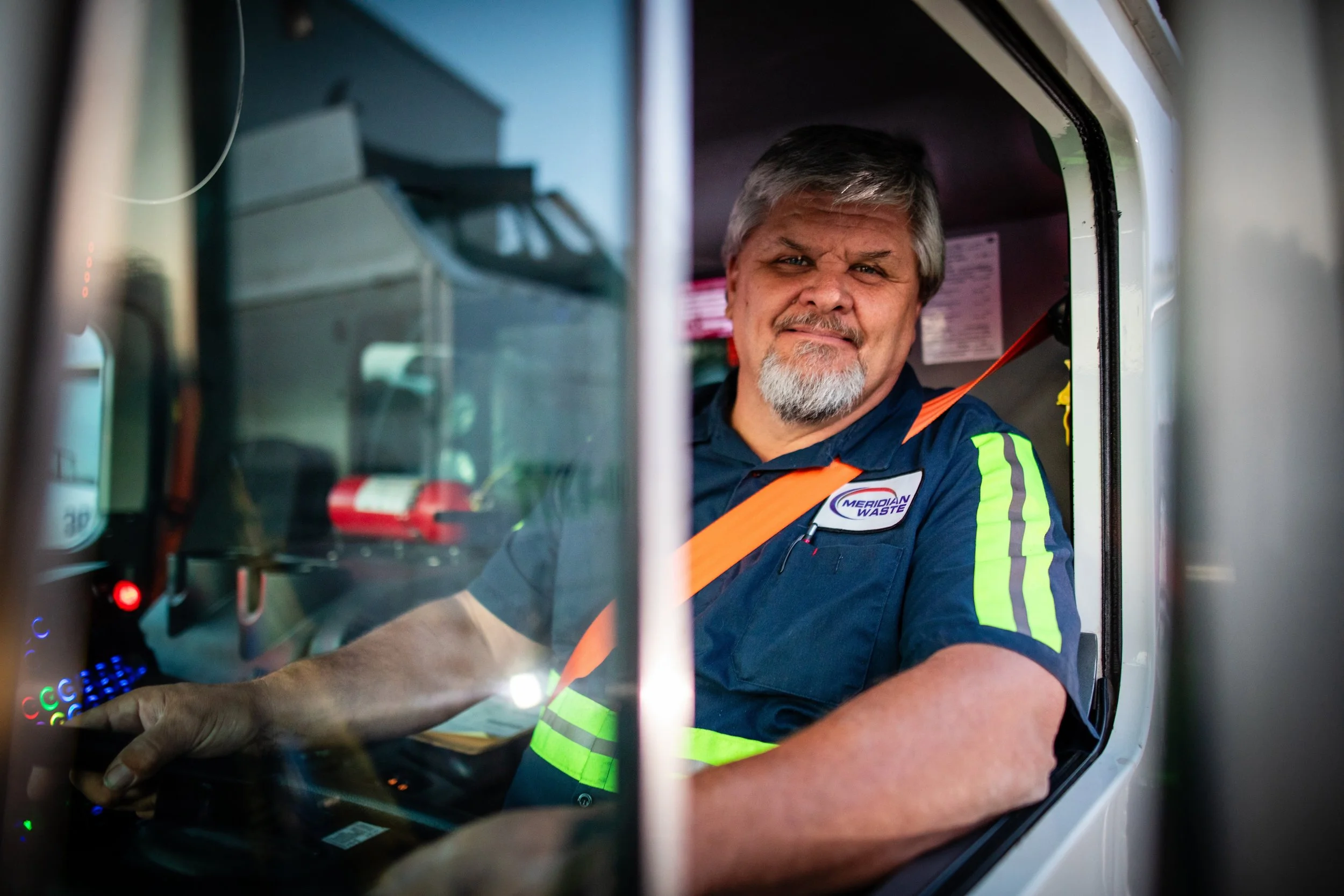 A waste management worker sitting in the driver's seat of a waste collection truck, wearing a navy blue uniform with reflective green stripes and a badge that reads 'Meridian Waste'.