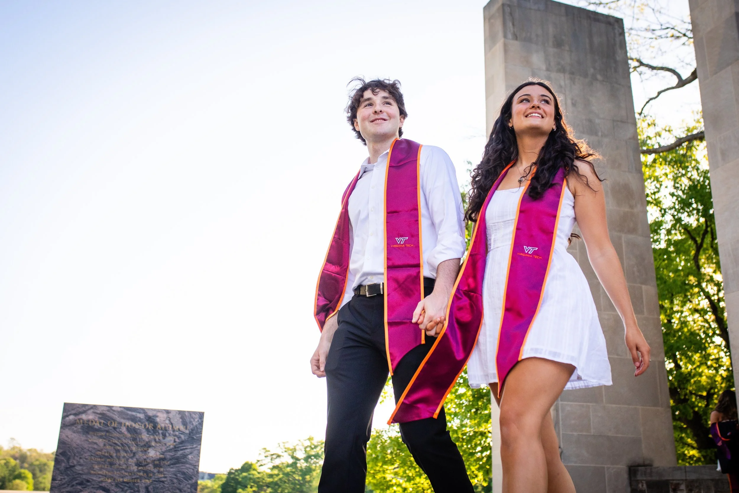 Two young adults, a man and a woman, holding hands and walking outdoors during a graduation ceremony, wearing purple academic stoles with the Virginia Tech logo, with trees and a monument visible in the background.