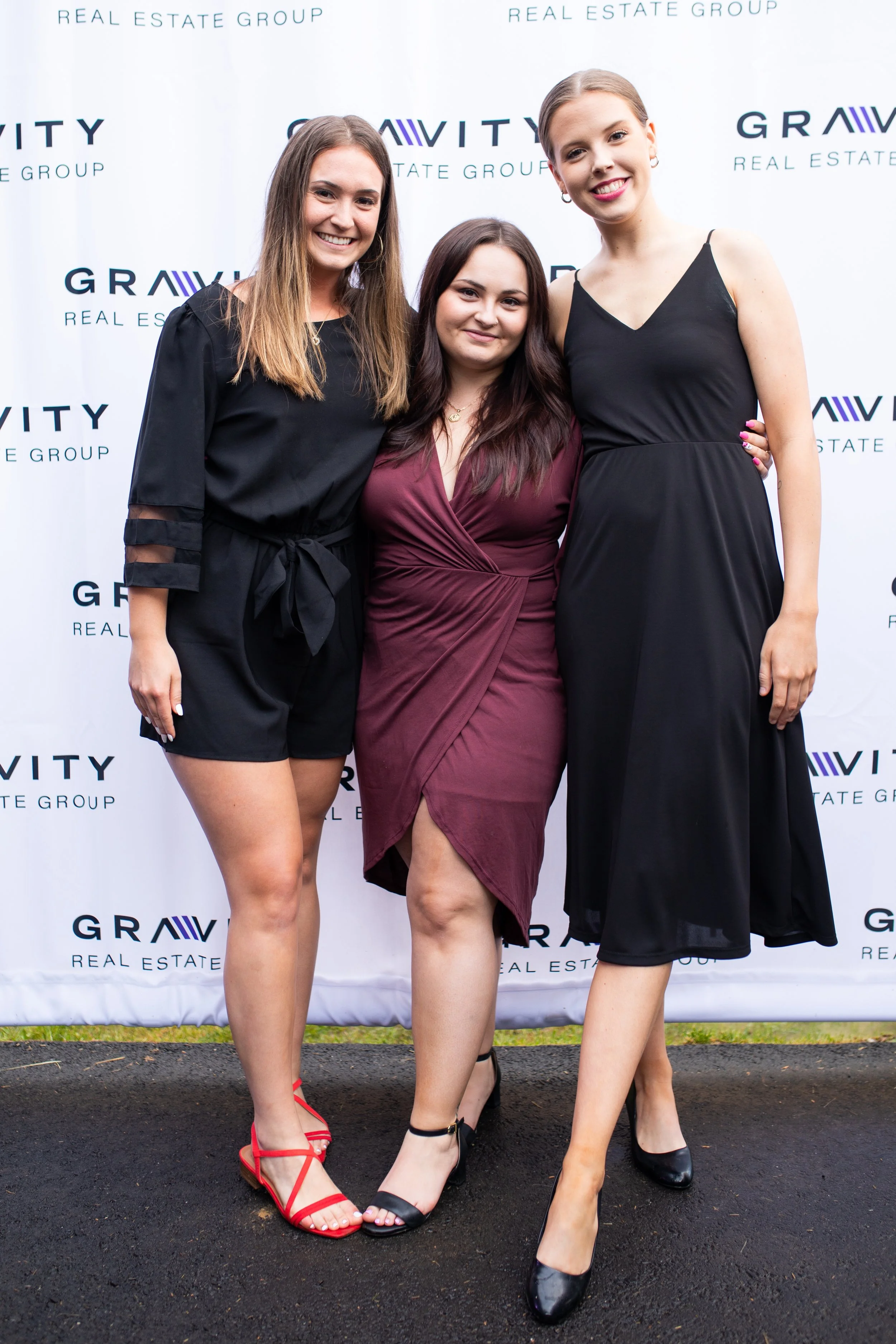 Three women smiling and standing together at an event with a backdrop that reads 'Gravity Real Estate Group'.