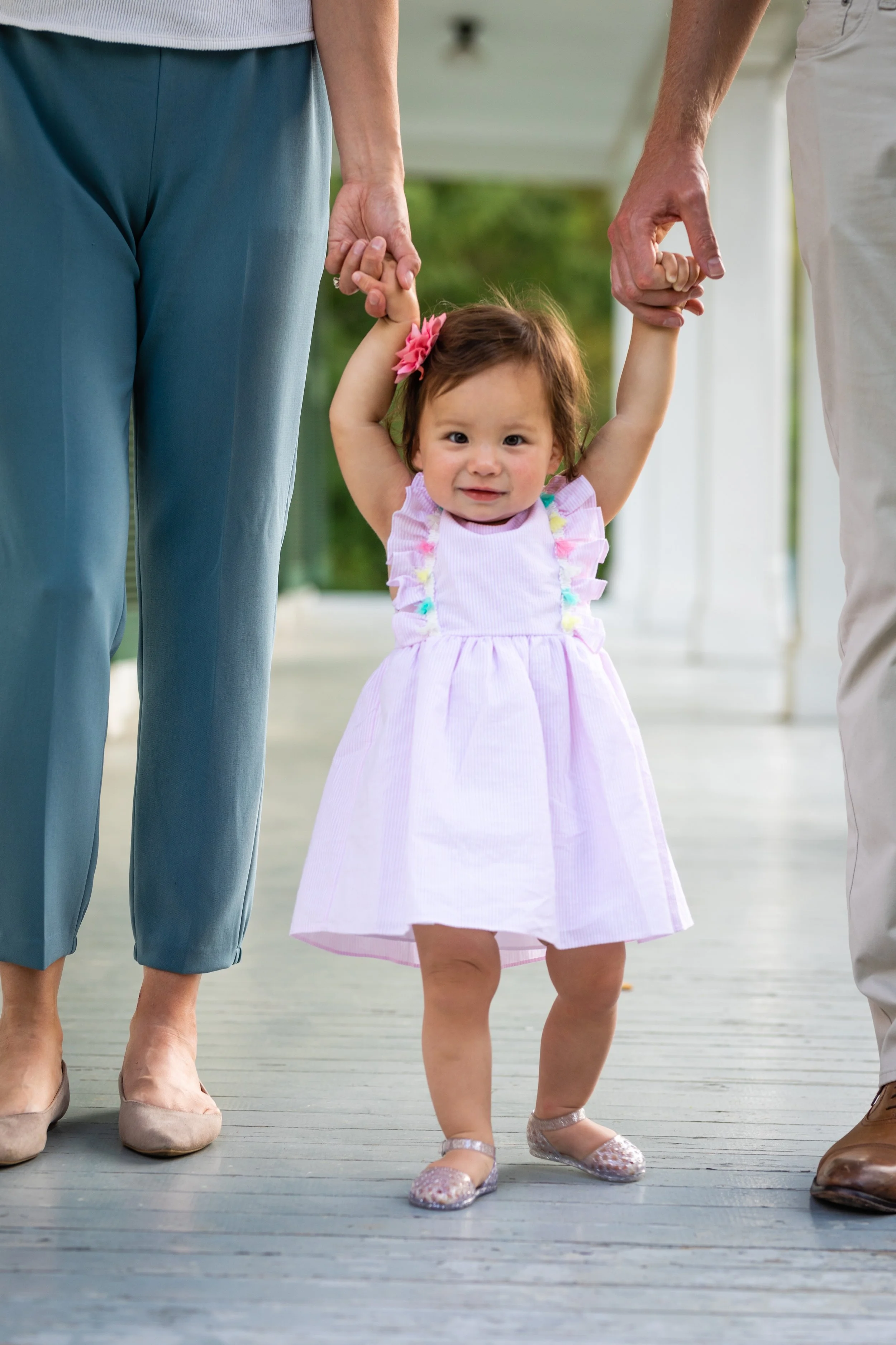 A young girl in a pink and white dress being supported by two adults as she walks on a porch, holding their hands with a worried expression.