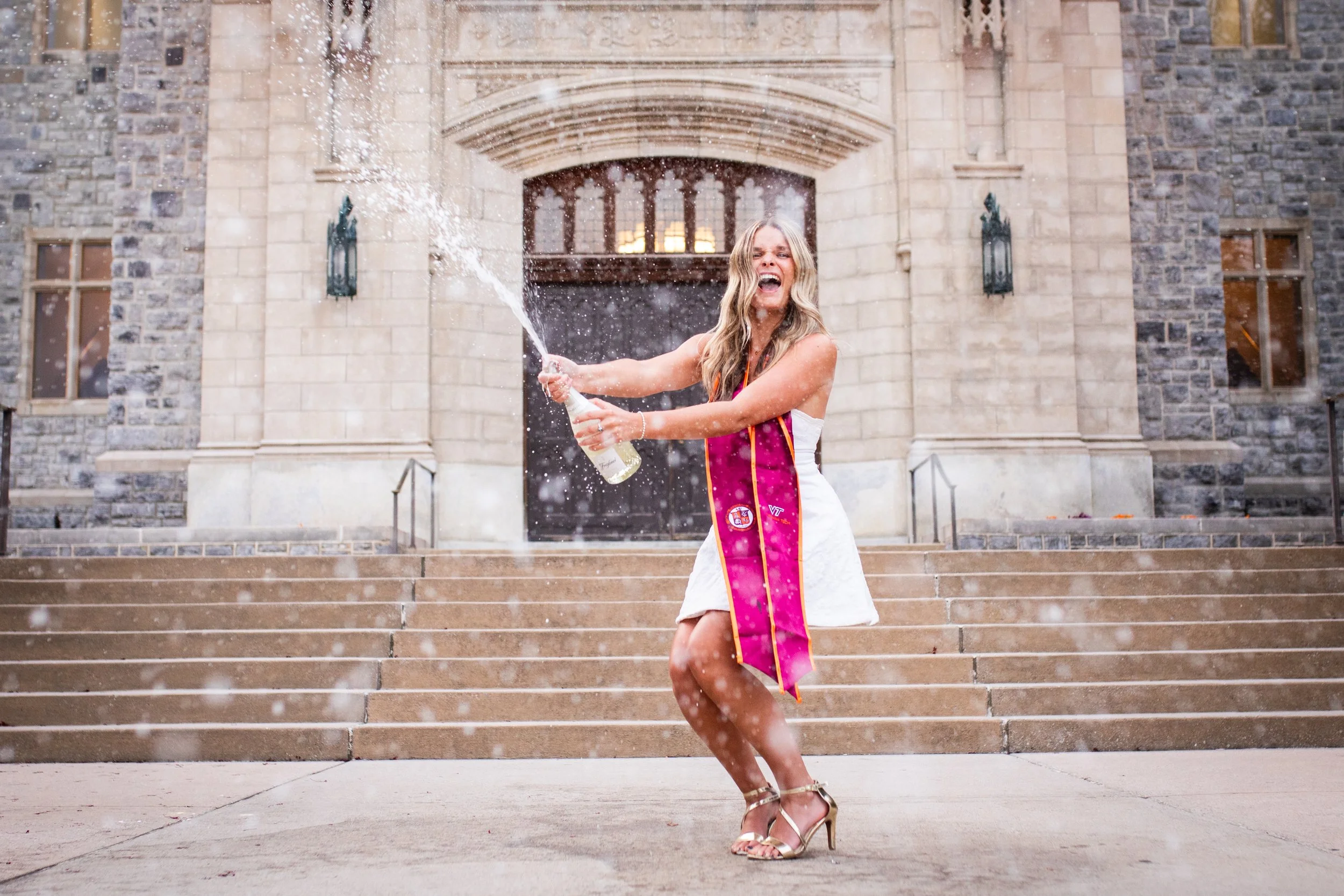 A woman in a white dress and high heels celebrating with a champagne bottle on the steps of a stone building, spraying champagne.