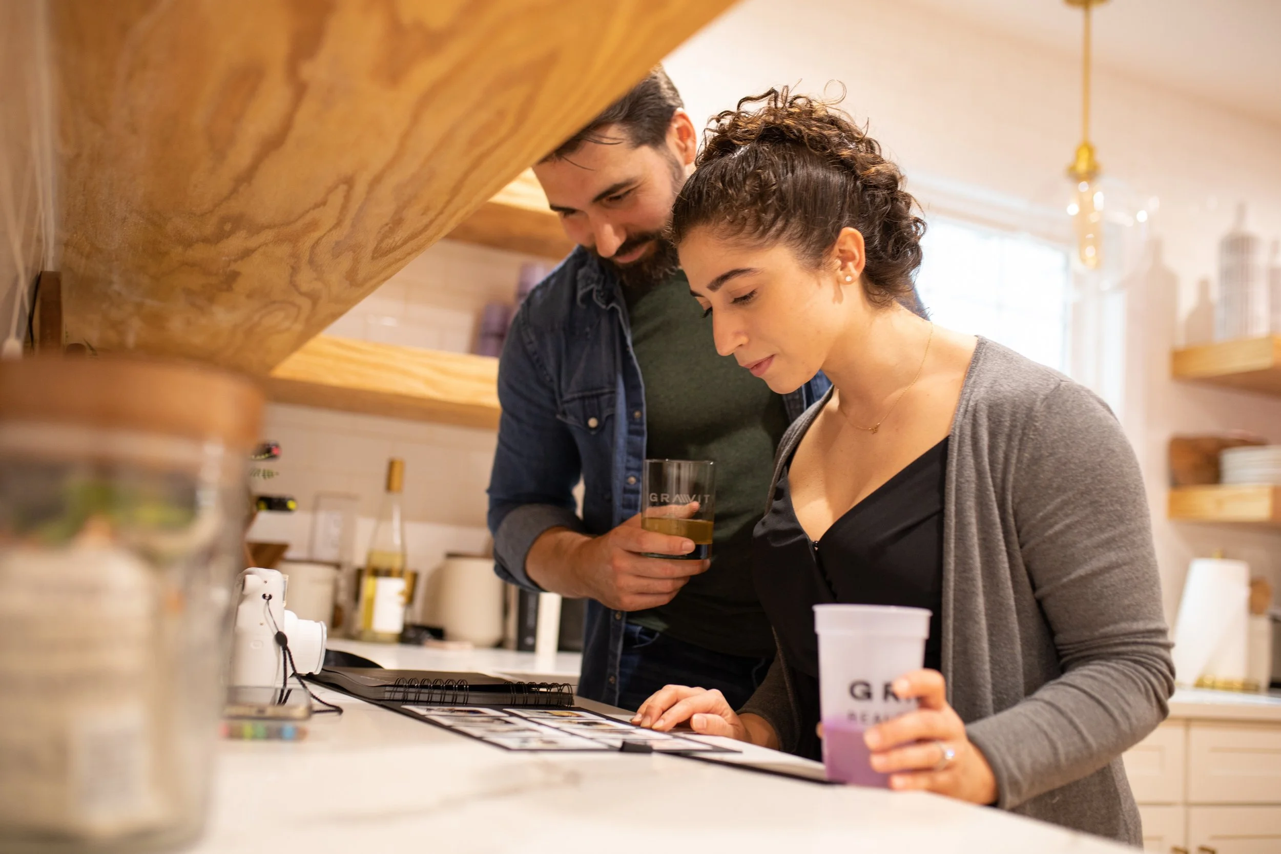A man and woman in a cozy kitchen looking at some documents or photos on the counter, with a drink in the man's hand and the woman holding a cup.