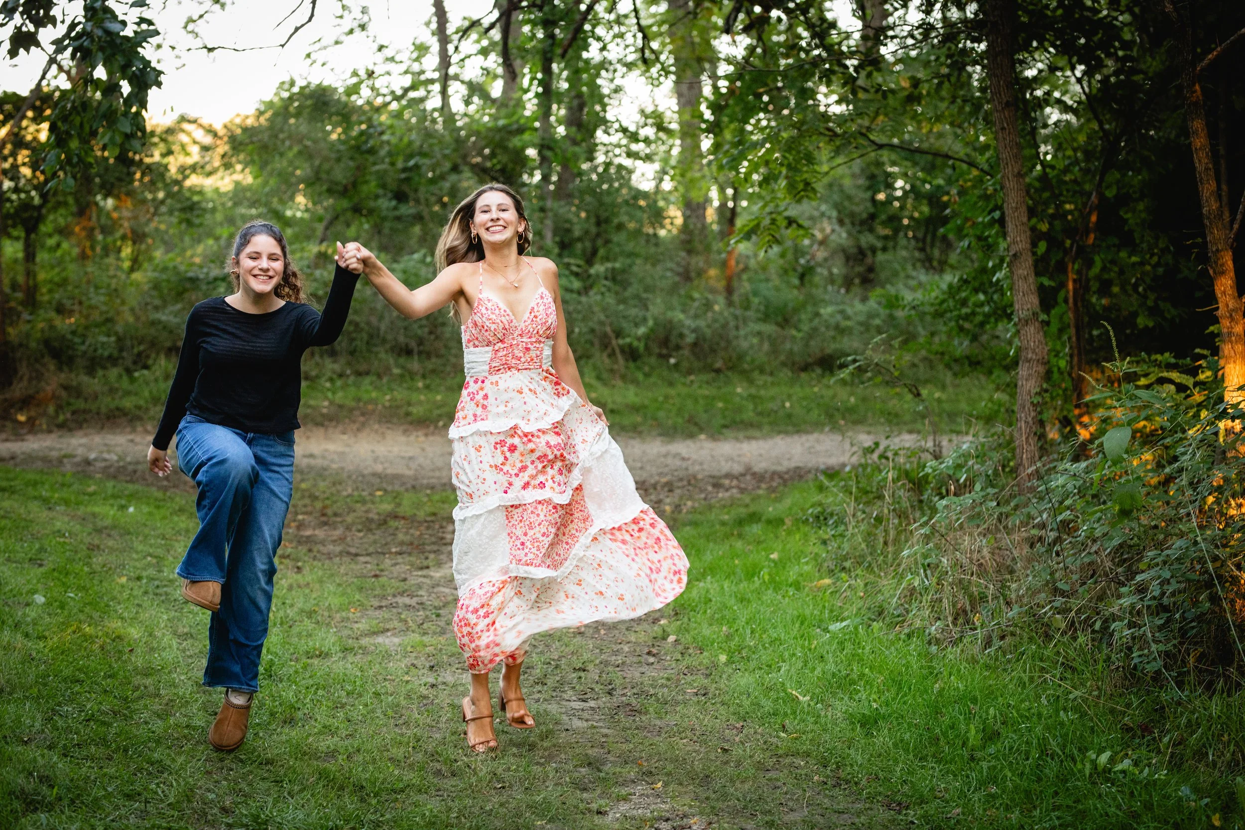 Two women are running and holding hands in a lush green forested area with sunlight filtering through the trees.