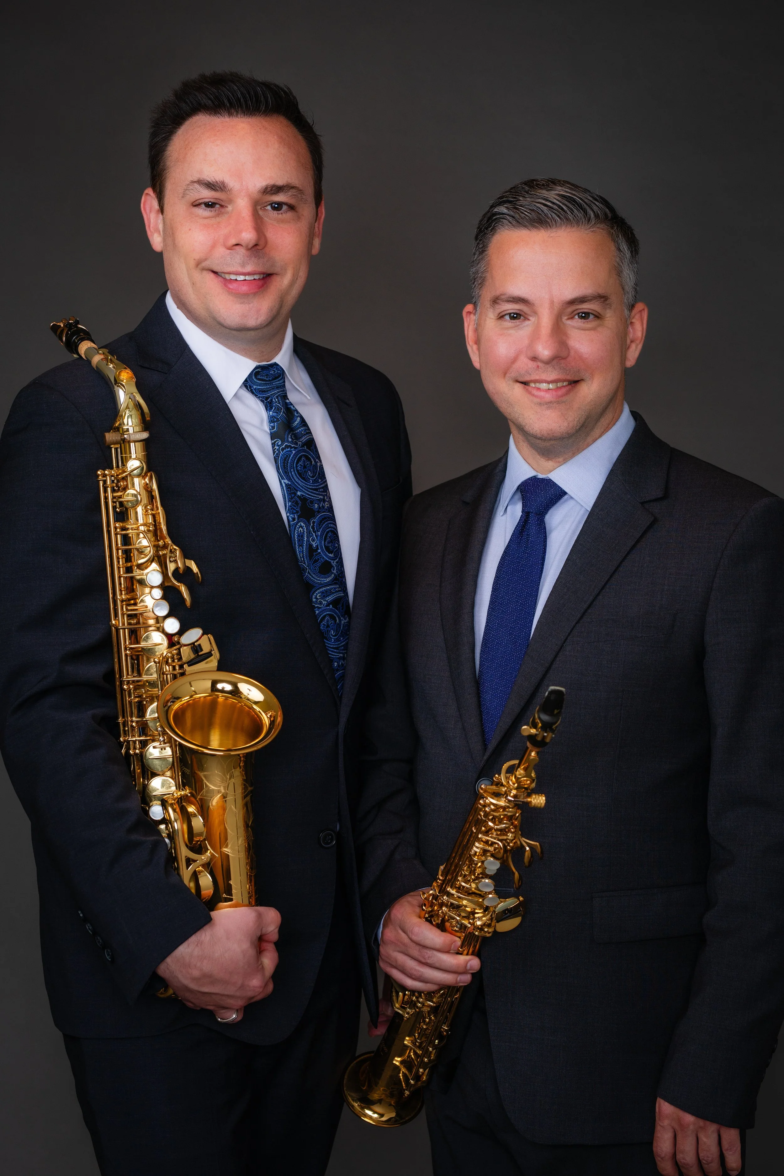 Two men dressed in formal suits holding saxophones, standing on a dark background and smiling at the camera.
