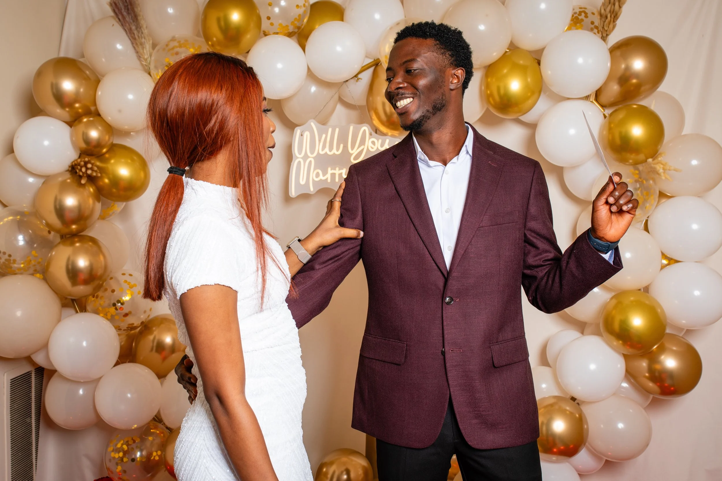 A man in a maroon suit and a woman with red hair in a white dress at a proposal event with balloon decorations and a sign that reads 'Will You Marry Me' in the background.