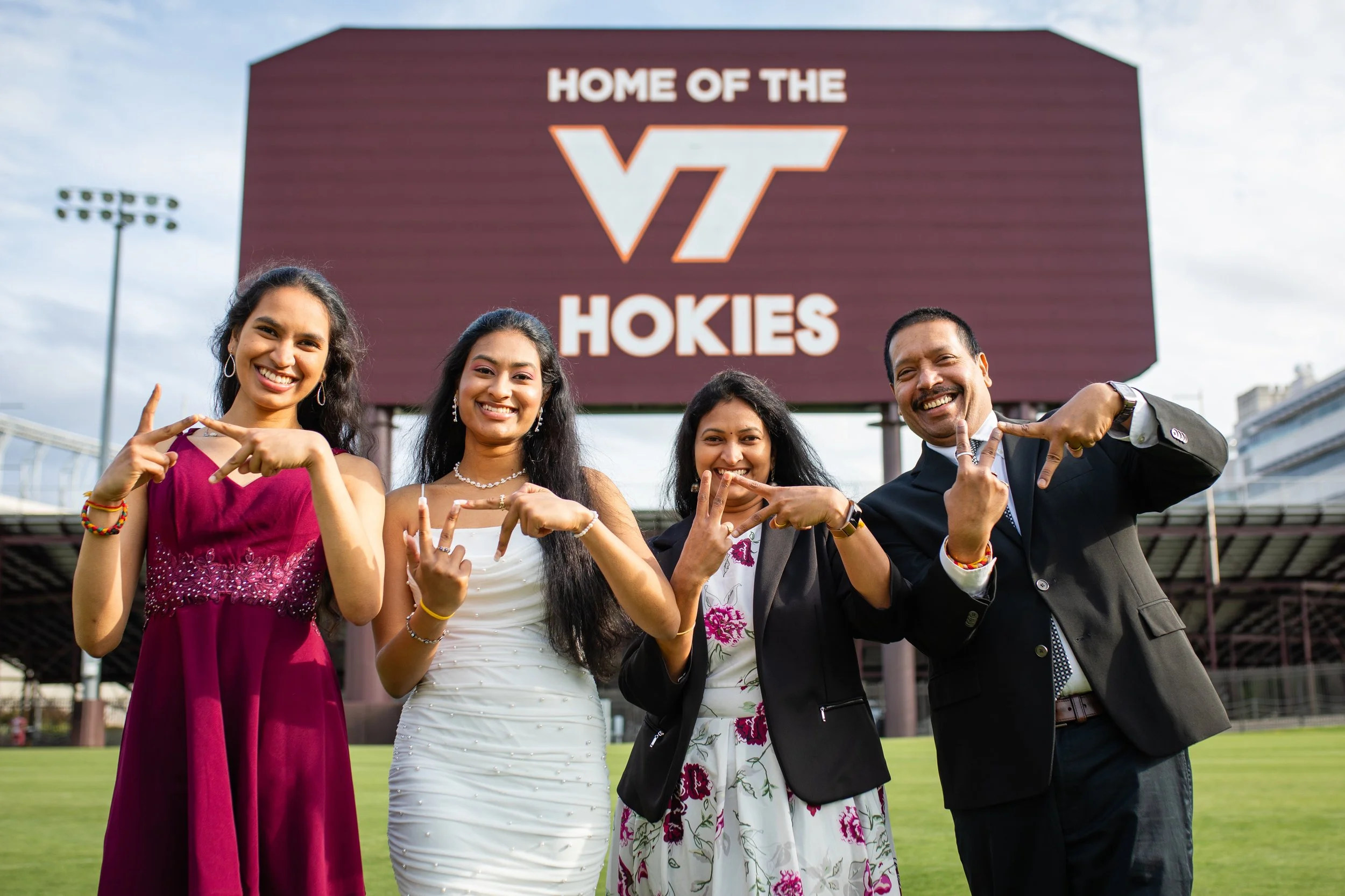Four diverse smiling people standing outdoors in front of a large sign that reads "Home of the VT Hokies," indicating they are at Virginia Tech. They are making heart and peace hand gestures and dressed in formal attire.