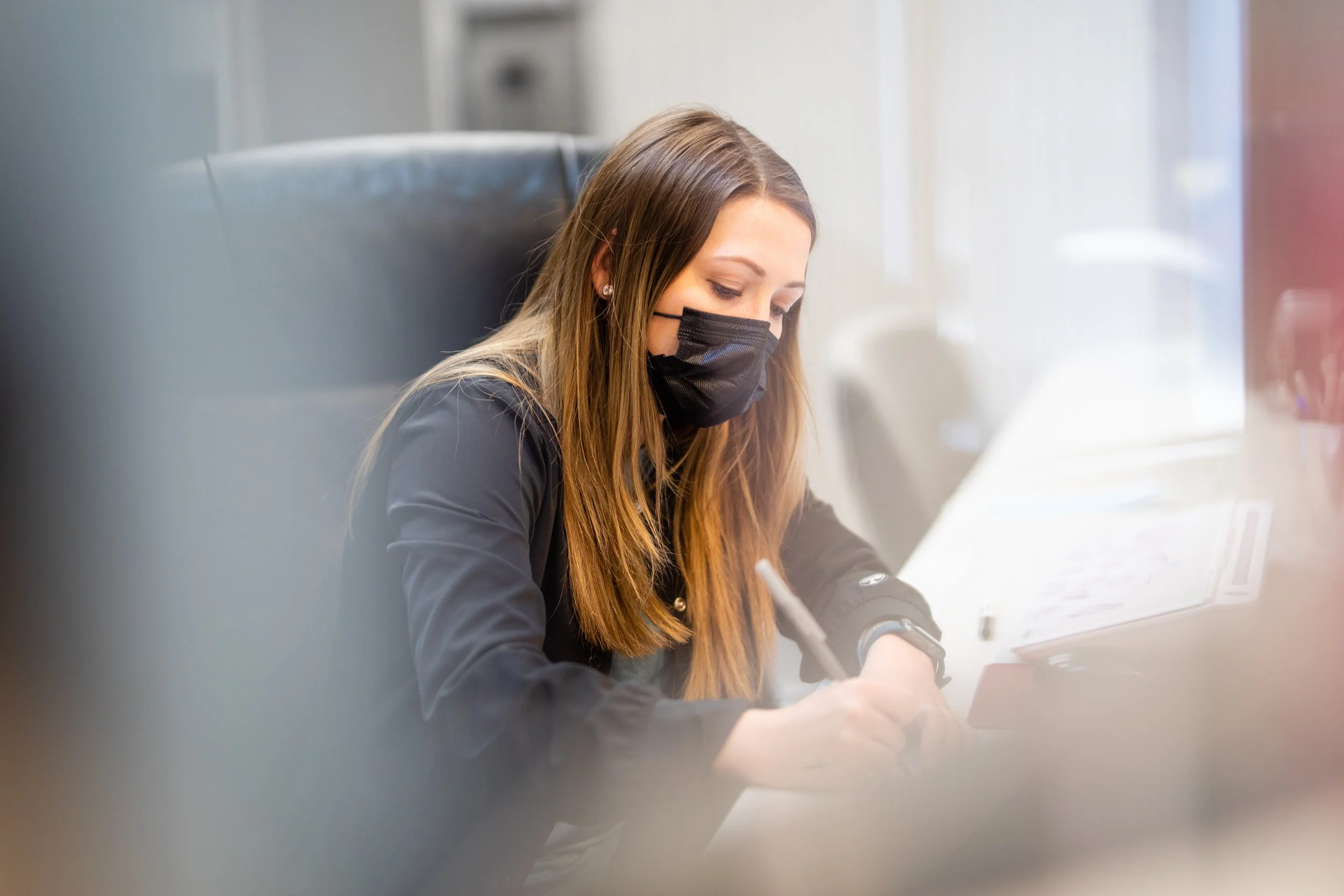 A woman with long brown hair wearing a black face mask and black jacket, sitting at a desk, writing with a pen on paper, in an office setting.
