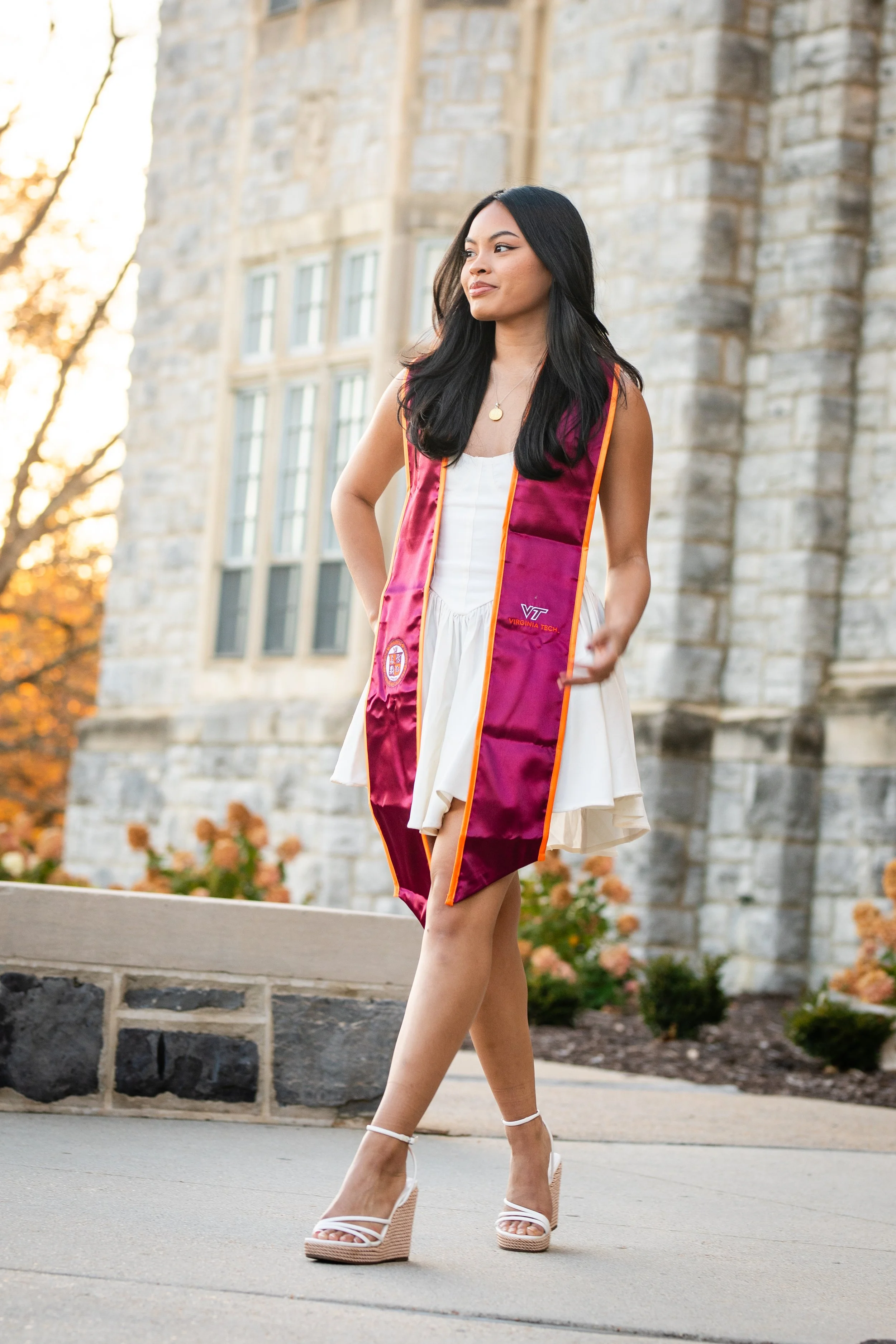 A young woman in a white dress and high heels is wearing a maroon graduation stole and walking outside on a college campus with a stone building and autumn trees in the background.