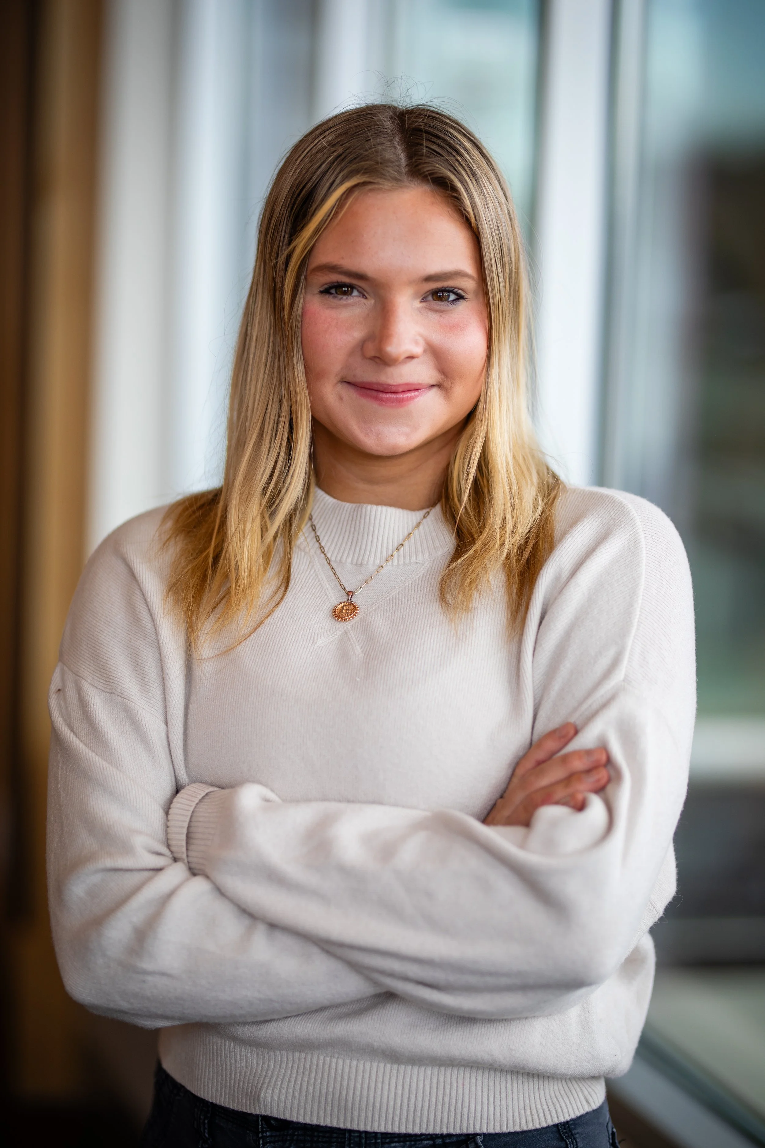 A young woman with blond hair wearing a white sweater and a necklace, standing indoors with her arms crossed, smiling at the camera.