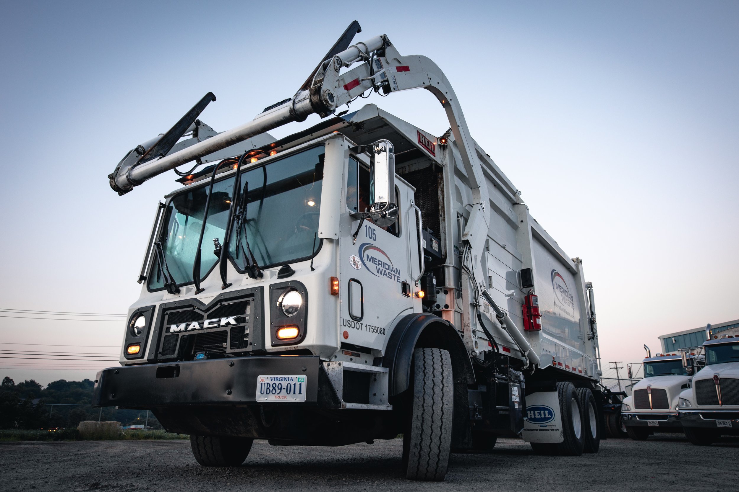 A white Meridian Waste trash collection truck parked on a dusty lot at dusk, with a blue sky background and some other trucks nearby.