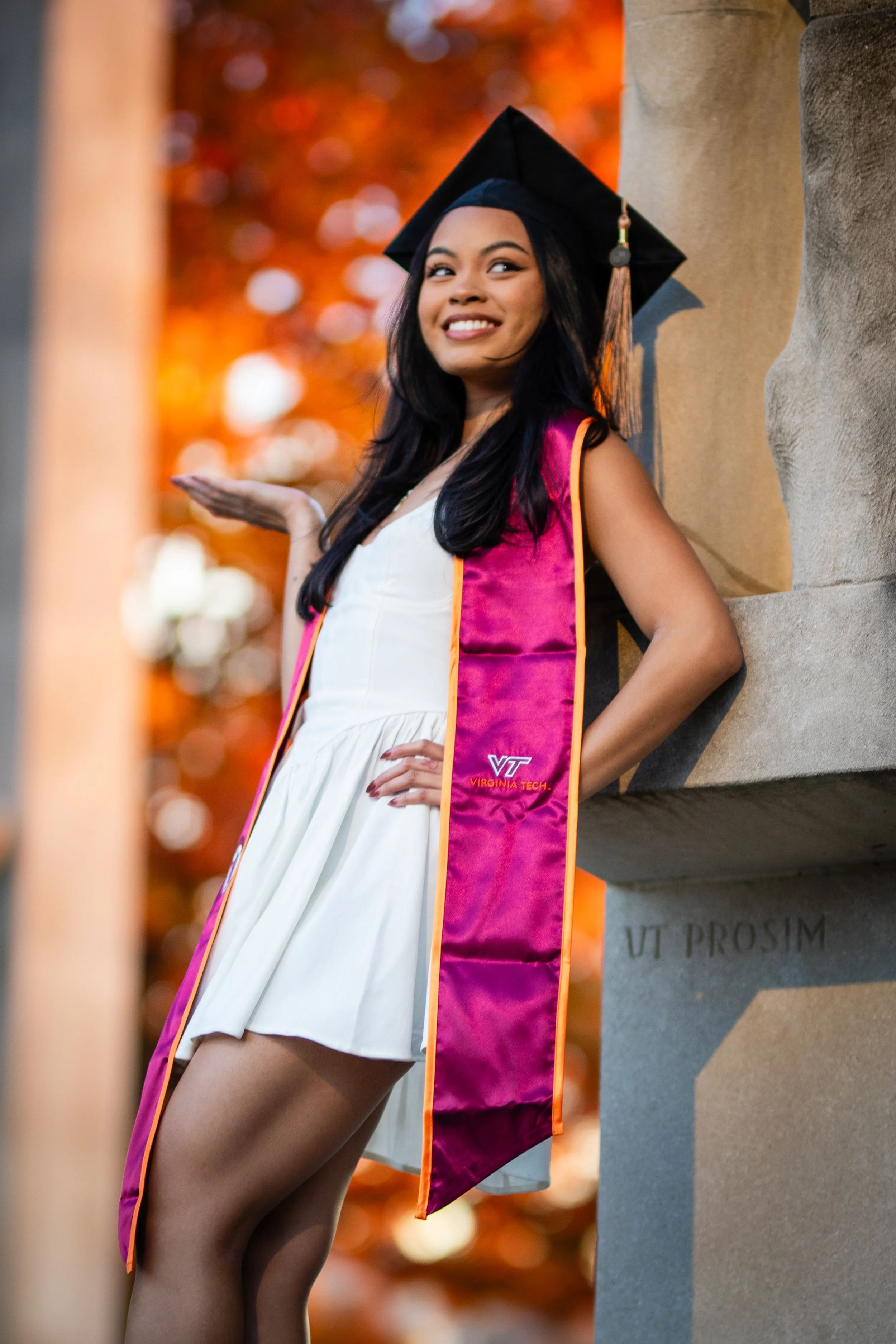 A young woman in a white dress and a black graduation cap with a pink and orange stole, smiling and posing outdoors during autumn.