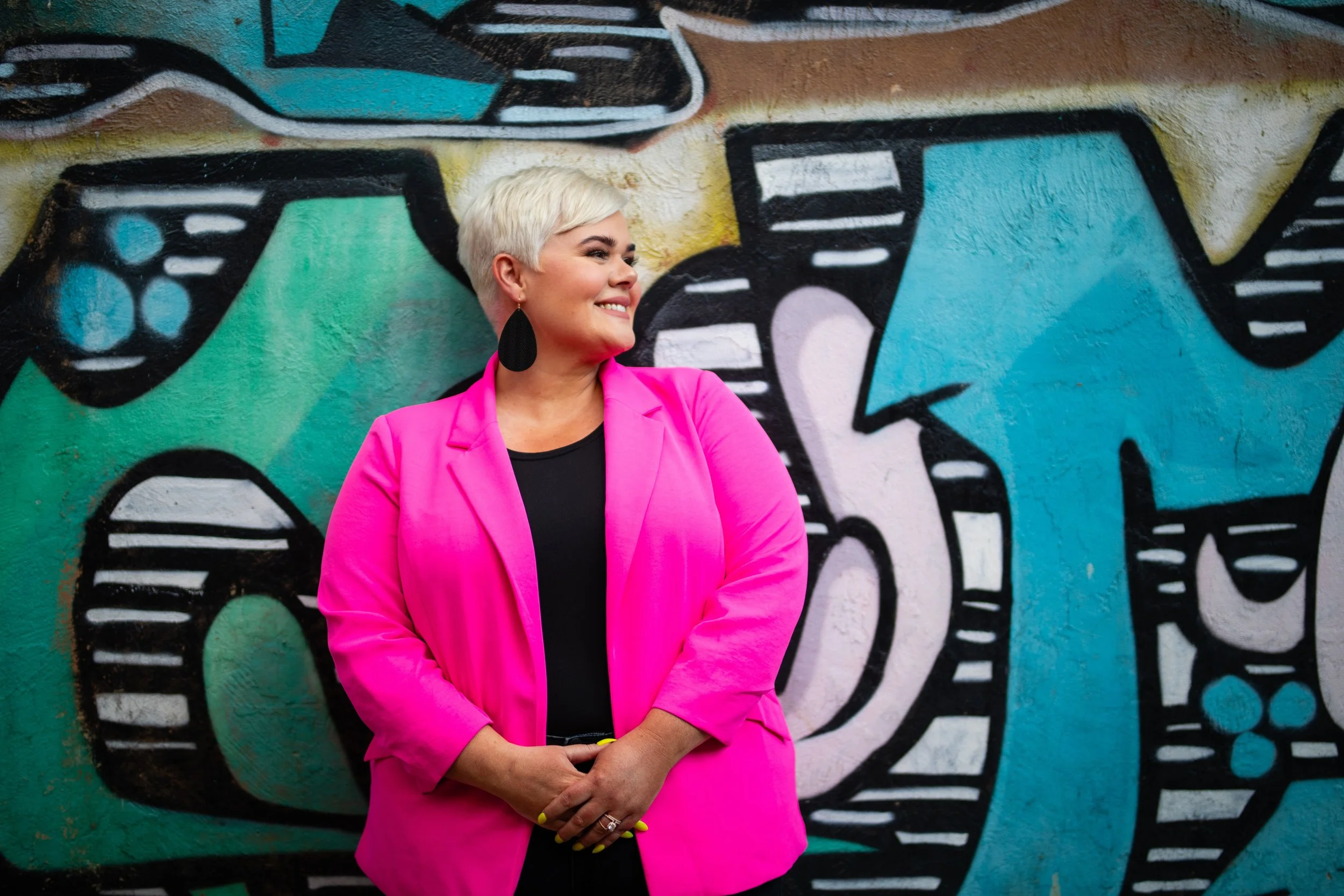Woman with short platinum blonde hair wearing a bright pink blazer and large black earrings, smiling and looking to her left, standing in front of a colorful graffiti wall with teal, black, and white abstract designs.
