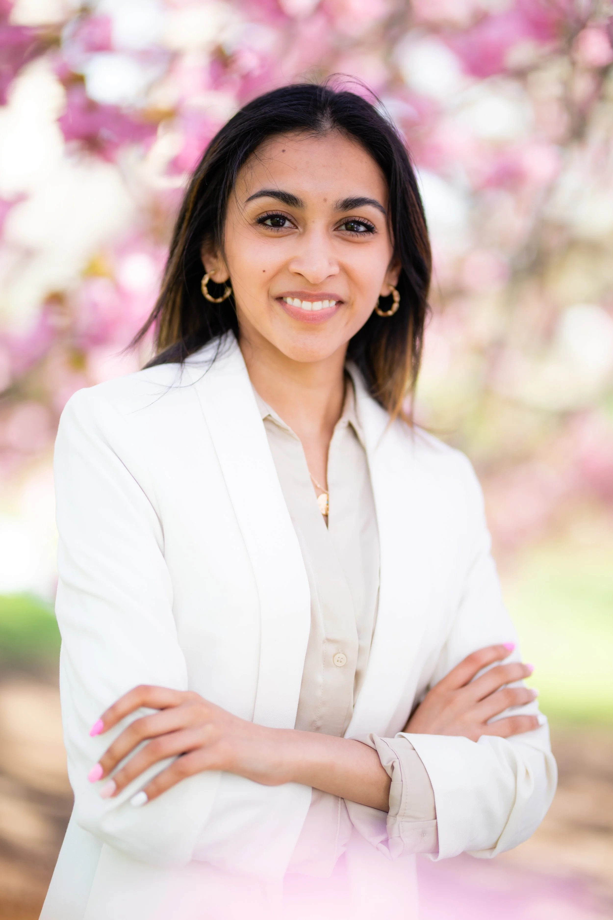 A woman in a white blazer standing outdoors with pink flowering trees in the background, smiling with arms crossed.