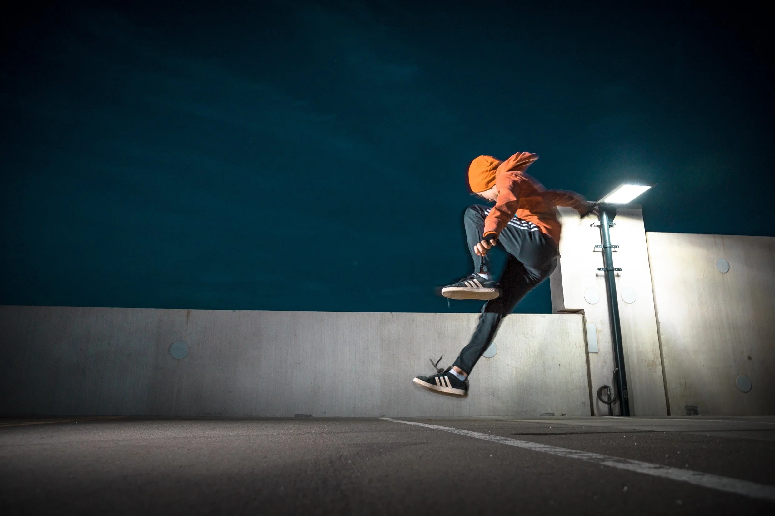 Person jumping in a parking lot at night, wearing an orange hoodie, black pants, and Adidas sneakers, with a streetlamp illuminating the scene.
