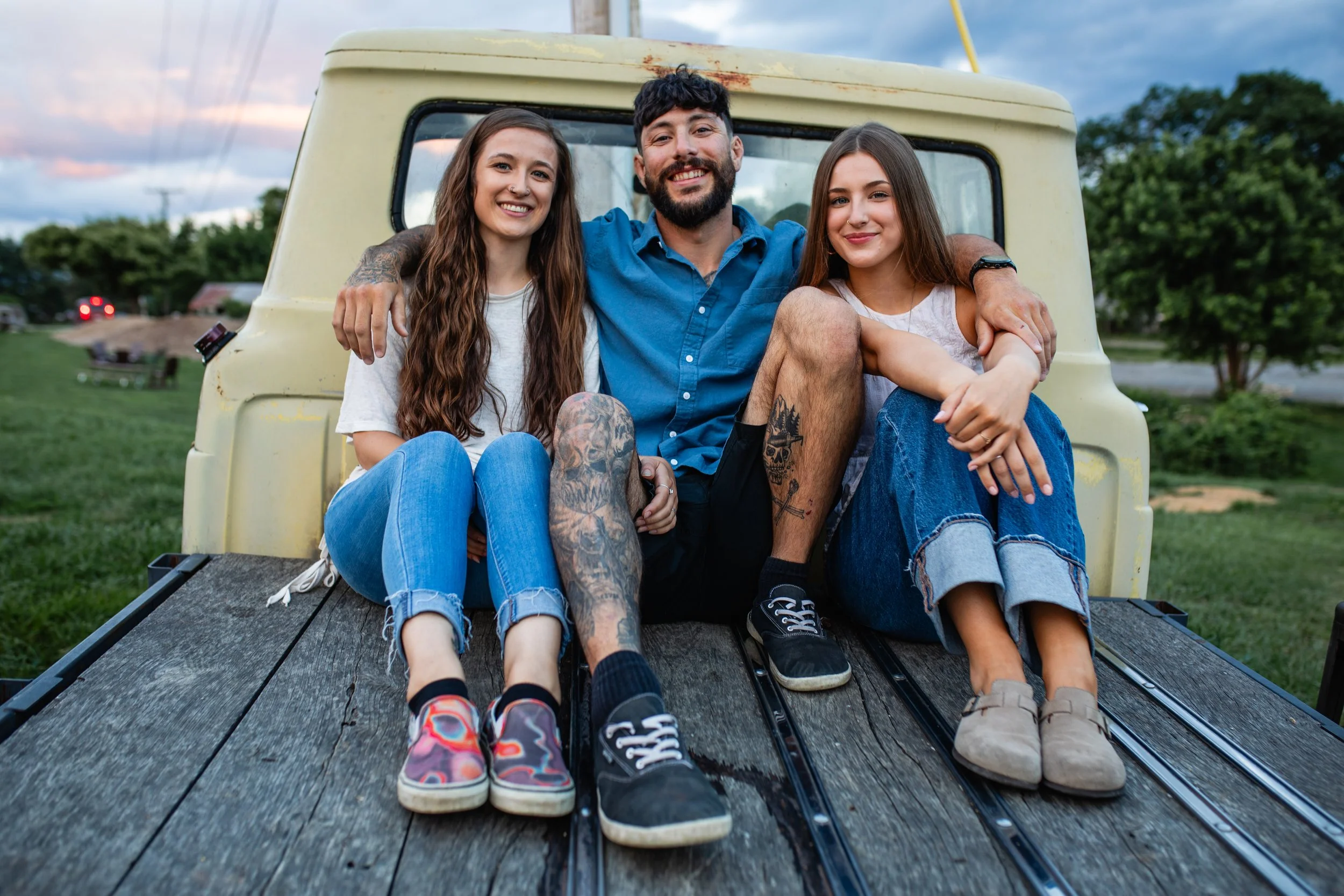 Three young adults sitting on the bed of a vintage truck outdoors during sunset, smiling and enjoying each other's company.