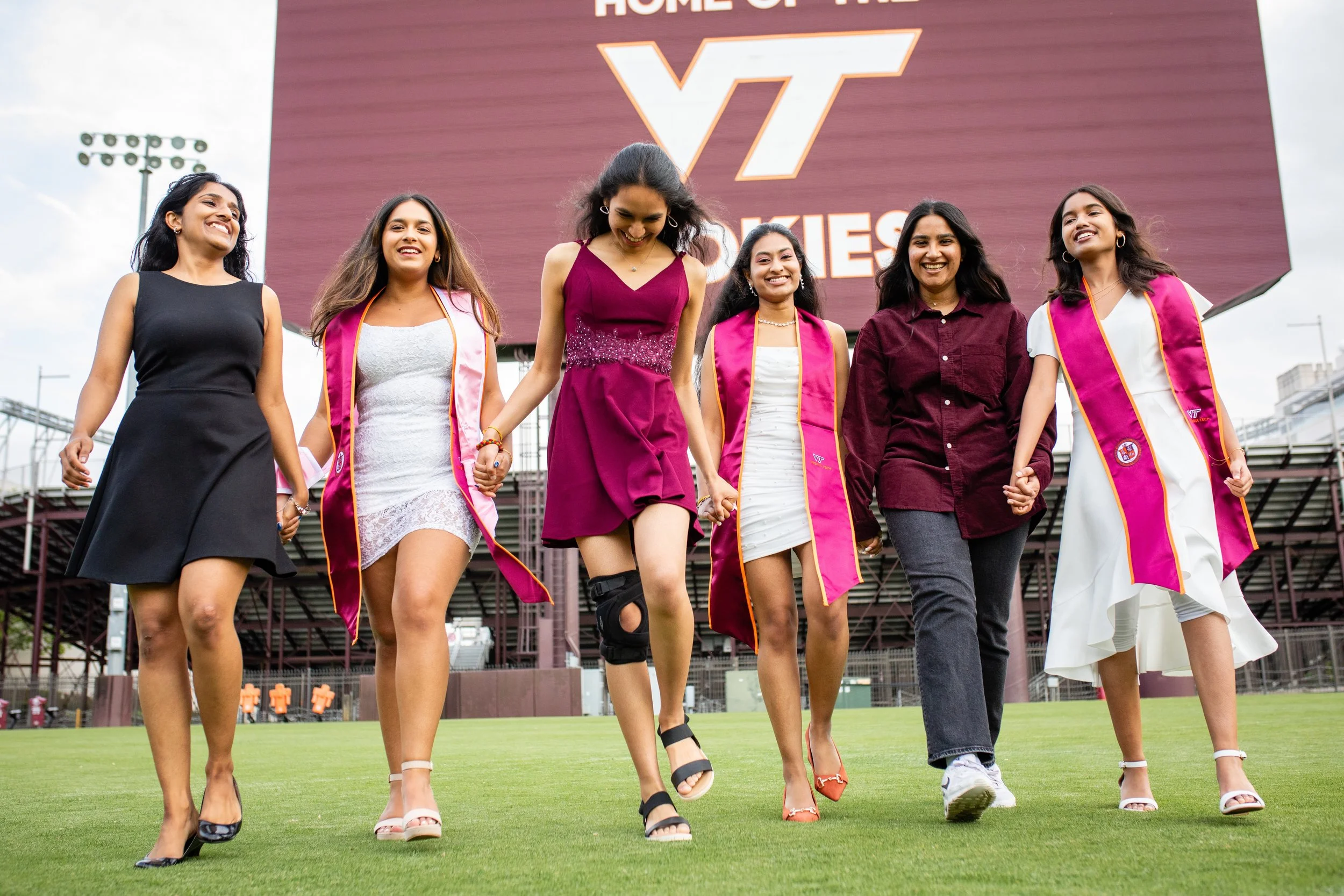 Group of six young women walking on a football field, some wearing graduation gowns with pink sashes, others in formal dresses, holding hands and smiling. Large scoreboard in the background with 'Home of VT' and 'Wahoos' text.