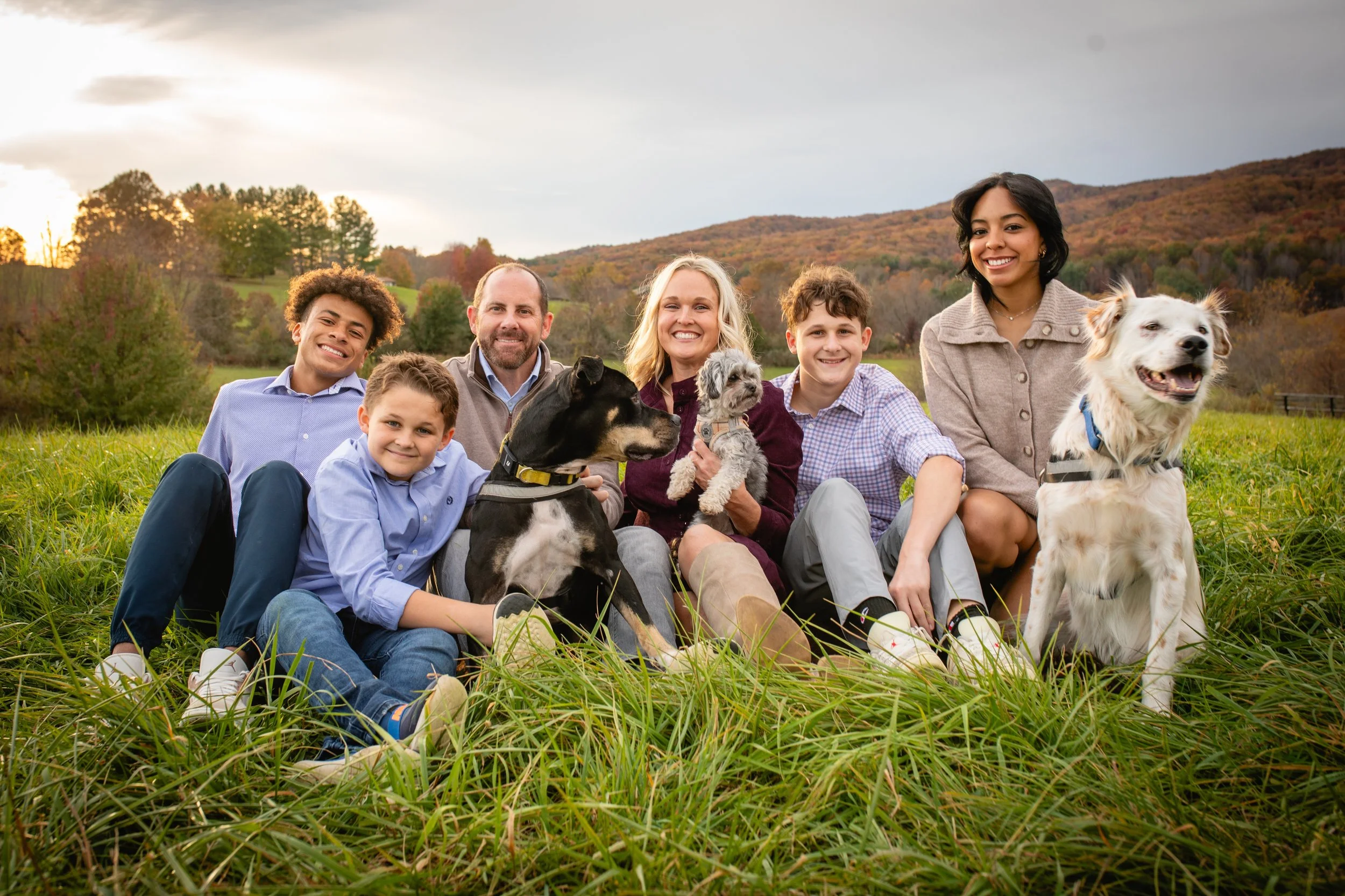 A family of seven people sitting on grass in a scenic outdoor setting with mountains and trees, smiling, with two dogs and two small dogs, during a sunset or sunrise.