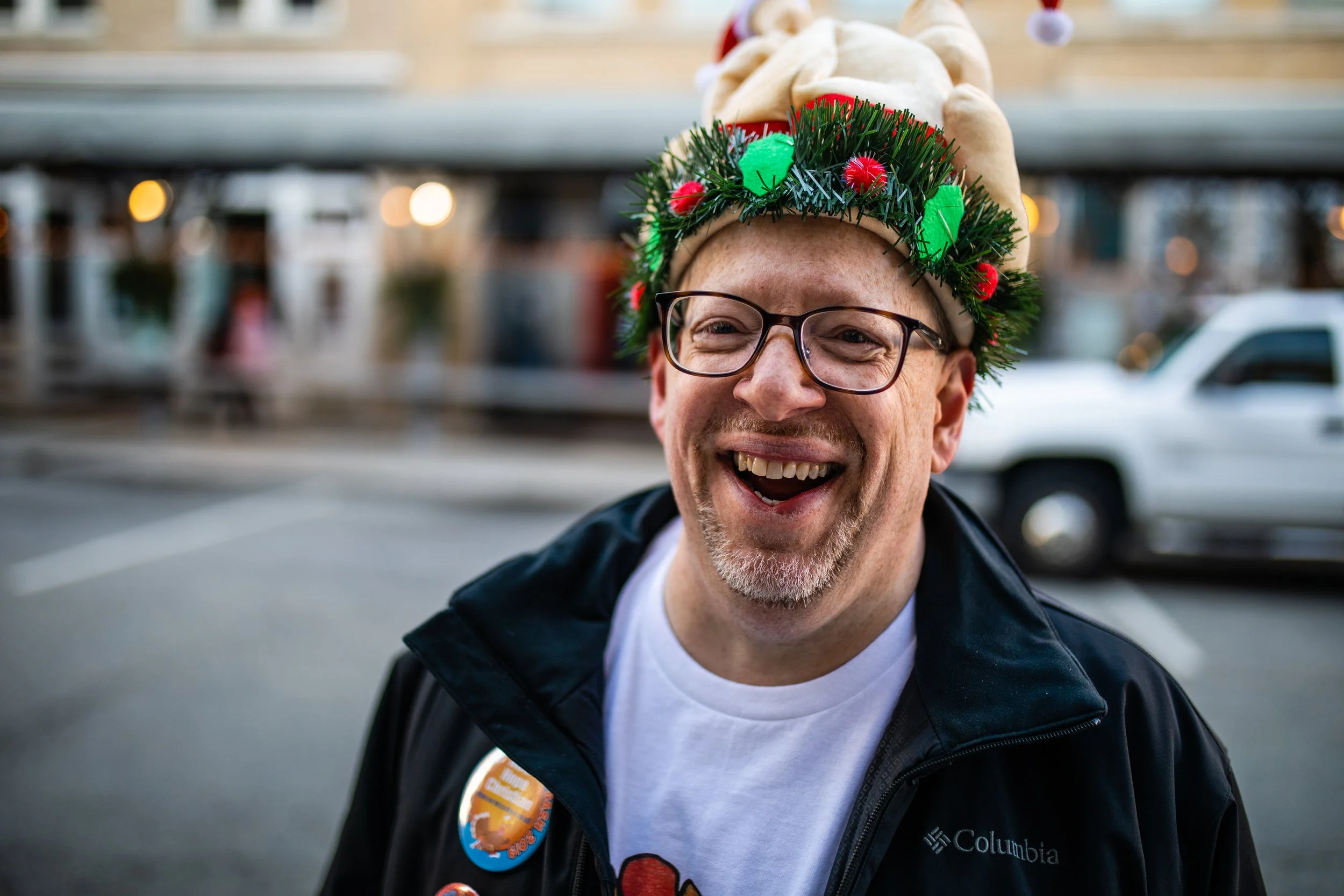 A smiling man wearing glasses, a Christmas-themed hat with plush parts, and a festive wreath around his hat. He's wearing a black Columbia jacket and standing outdoors with blurred cars and buildings in the background.